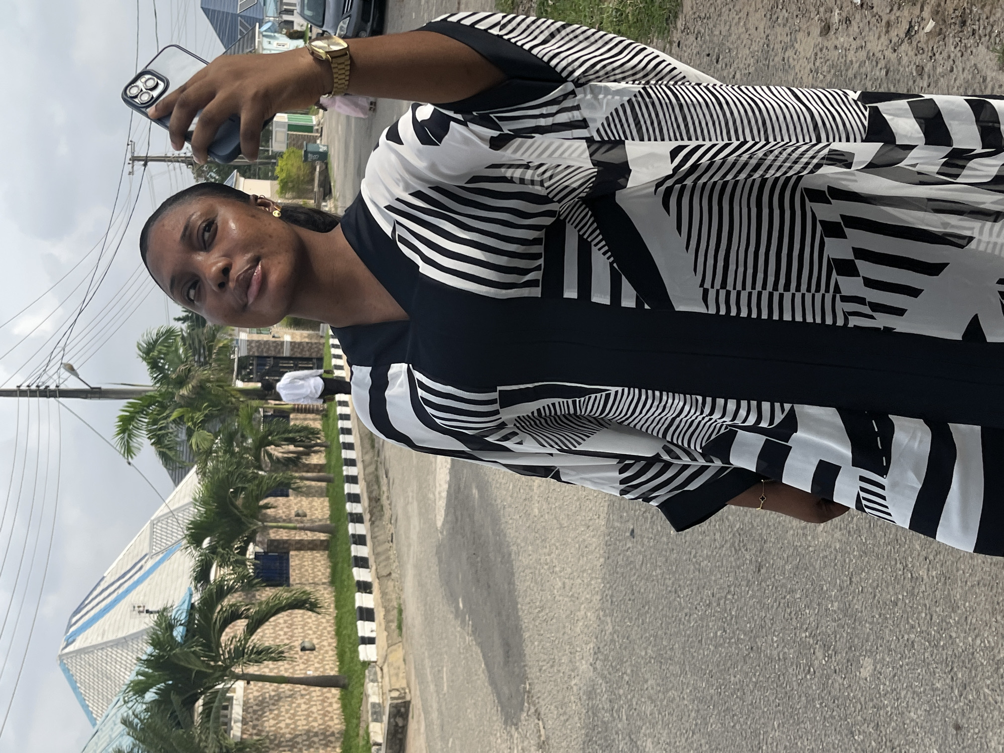 A woman is taking a selfie outdoors on a city street. She is wearing a black and white patterned dress and a gold watch. She is holding a smartphone in her left hand. Palm trees and buildings are visible in the background, with a modern building with a white exterior and blue trim being prominent. Power lines are strung overhead. The location appears to be in Abuja, Nigeria, given the style of architecture and landscaping.