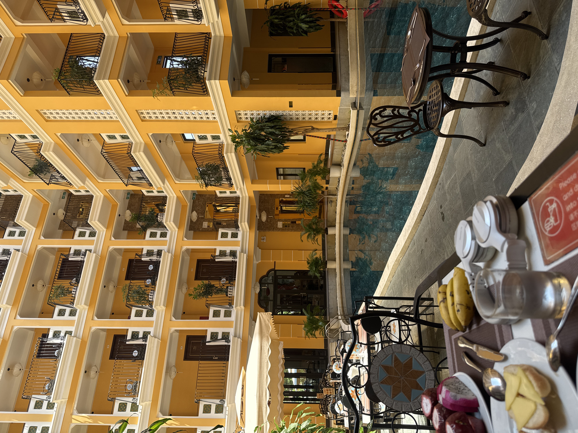 The image depicts a courtyard scene, possibly within a hotel or resort, in Đà Nẵng, Vietnam. The architecture is characterized by a multi-story structure with yellow walls, balconies adorned with wrought iron railings, and plants. The courtyard features a shallow water feature with reflective qualities, visible tables and chairs, and plants. A table holding a breakfast or brunch setup, including fruit (bananas, dragon fruit), and dishes. No individuals are clearly visible. The scene appears to be a daytime setting.