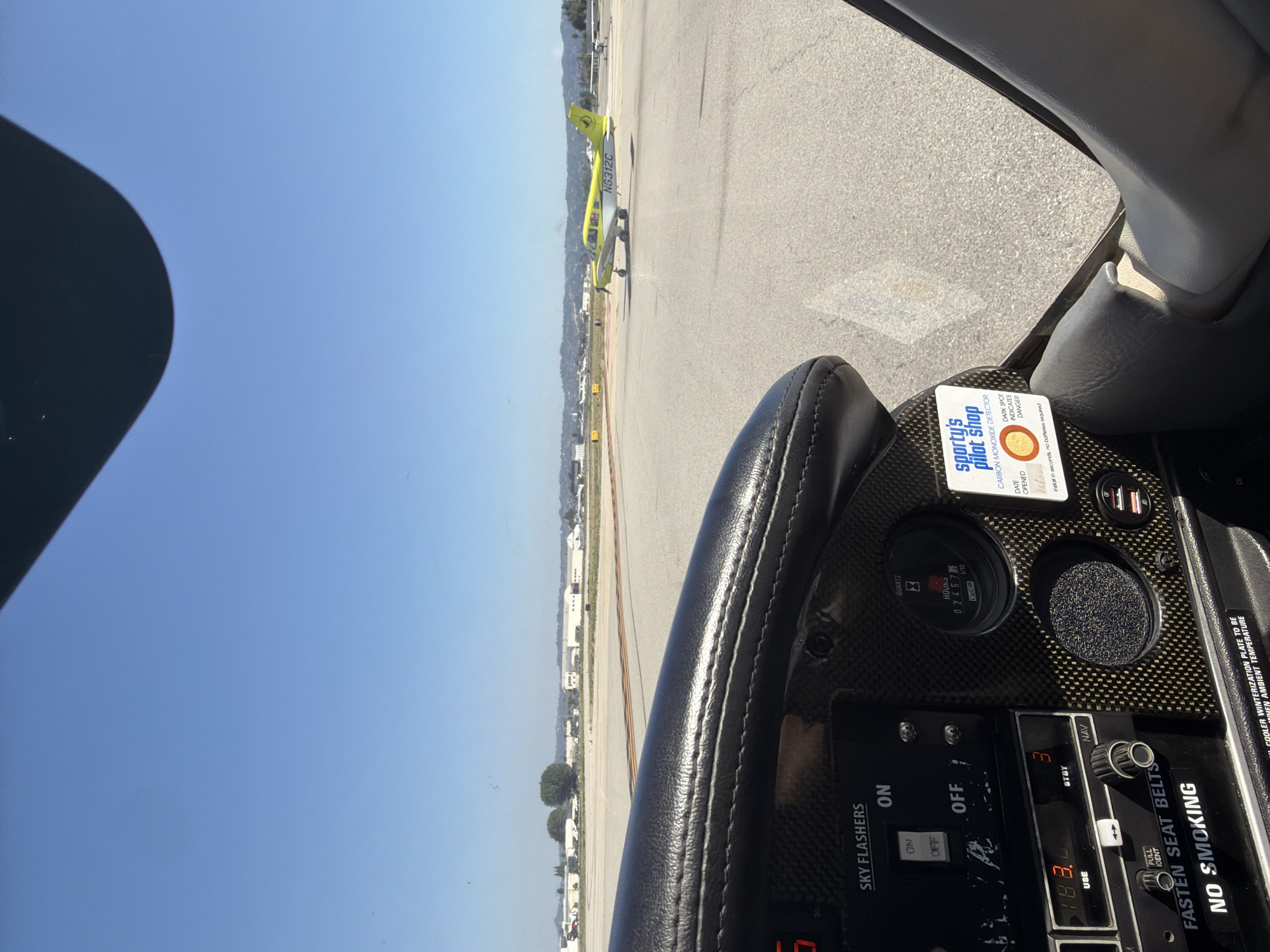 The image shows the view from the cockpit of a small airplane, likely on the ground at an airport. Visible through the windshield is another airplane, painted yellow and white, with the tail number "N6312C". The cockpit instrumentation panel is detailed, displaying switches, gauges, and warning labels such as "FASTEN SEAT BELTS" and "NO SMOKING." A "Sporty's Pilot Shop" carbon monoxide detector sticker is affixed to the panel. The sky is clear and blue. Based on visual cues like the type of aircraft and airport infrastructure, the location is likely an airport within the city of Los Angeles, United States of America. The perspective is from inside one of the aircraft, focusing on the other aircraft at the airport.