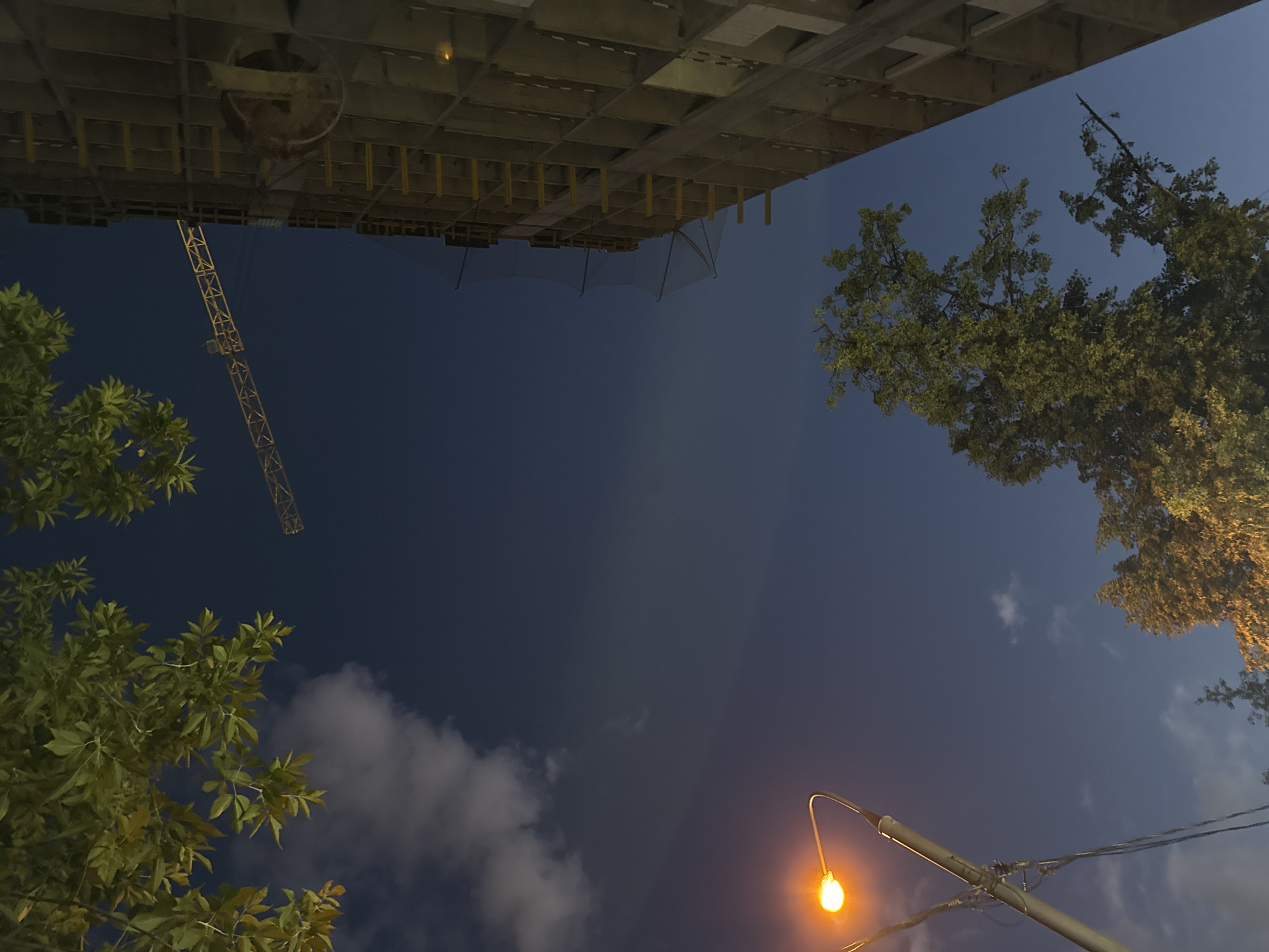 The image depicts a nighttime scene in Moscow, Russia, with a construction site as a prominent element. A tall, unfinished building occupies the right side of the frame, its skeletal structure of concrete and scaffolding illuminated by artificial light. A construction crane is visible extending from the building into the deep blue night sky, its metallic lattice stark against the darkness. In the foreground and upper left corner, the dark green leaves of a tree hang into the frame, their outlines softened by the low light. A single street lamp with a bright, orange glow is positioned in the lower left quadrant, casting a warm light on its pole and illuminating a section of the sky. Several wispy clouds are scattered across the sky, adding texture to the deep blue expanse. The overall impression is one of urban development under a twilight sky.