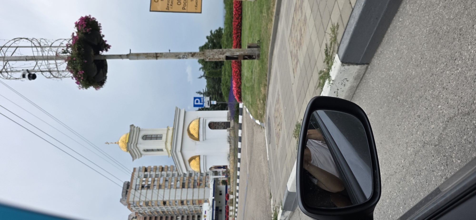 The image, taken in Kislovodsk, Russia, shows a small white chapel with golden domes, a tall building in the background, and a roadside with a traffic sign indicating parking. A side mirror of a car reflects a person wearing a white t-shirt. A lamppost with a basket of flowers and a security camera is also visible. The sky is overcast.