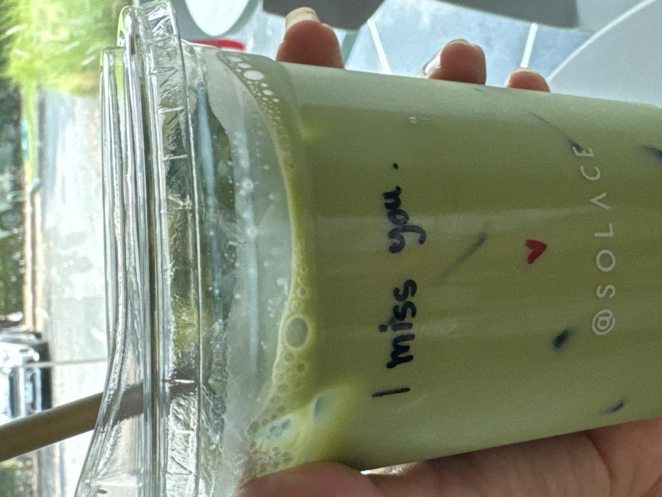 A translucent plastic cup of matcha latte with ice is held in someone's hands. The drink is a pale green color with foam at the top. Written on the side of the cup in black marker is the phrase "I miss you," with a small red heart drawn below. Printed vertically is "@SOLACE." The cup has a ridged lid with a drinking straw visible. A window is visible through the cup, showing greenery and a parked car.