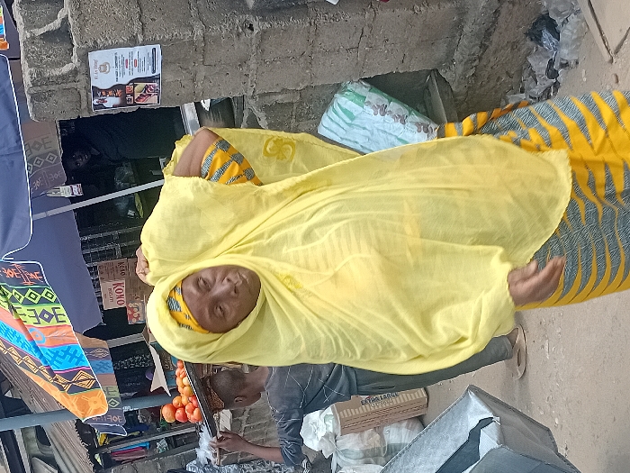 A woman wearing a yellow headscarf stands in a market setting. She has a patterned dress with yellow and grey-blue stripes. A man next to her hands her something while standing beside a display of oranges. Nearby are various goods, including fabrics with colorful patterns and boxes, positioned within what appears to be a bustling commercial area.