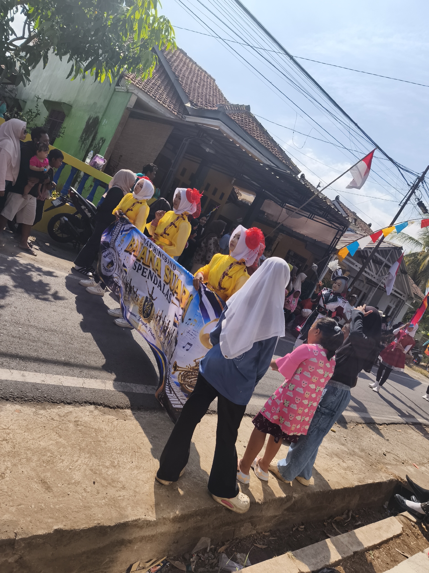 In Kutoarjo, Indonesia, a marching band parade is underway on a sunny day. The focus is on a group of young people in colorful costumes. Two girls in yellow uniforms with red headpieces and white hijabs are prominently featured, carrying a banner that reads "MARCHINGBAND DANA SUARA SPENDALAS". The banner also depicts musical instruments and a stylized image of a marching band. Several other individuals are visible, including spectators and participants. The backdrop consists of houses, trees, and overhead utility wires. Decorative flags are strung across the street, adding to the festive atmosphere. The ground is paved with asphalt and concrete, and a storm drain is visible along the side of the road.