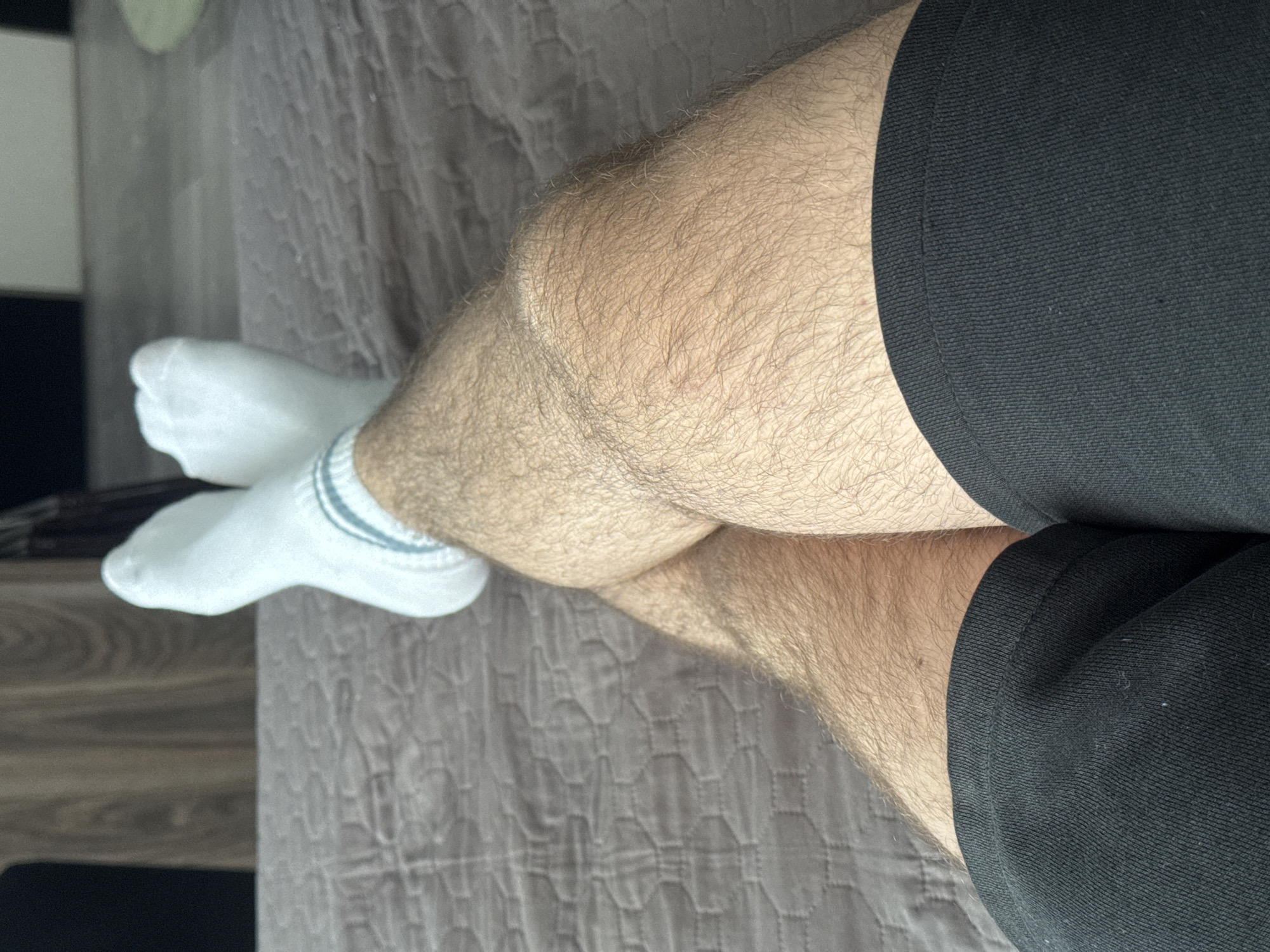 A close-up shot shows a person sitting with their legs crossed. They are wearing black shorts and white socks with grey stripes. The legs are hairy. The background consists of a grey, patterned surface, possibly a bedspread or a quilt. Part of a dark-colored object is visible in the upper left of the shot. The location is not discernible from the image alone.
