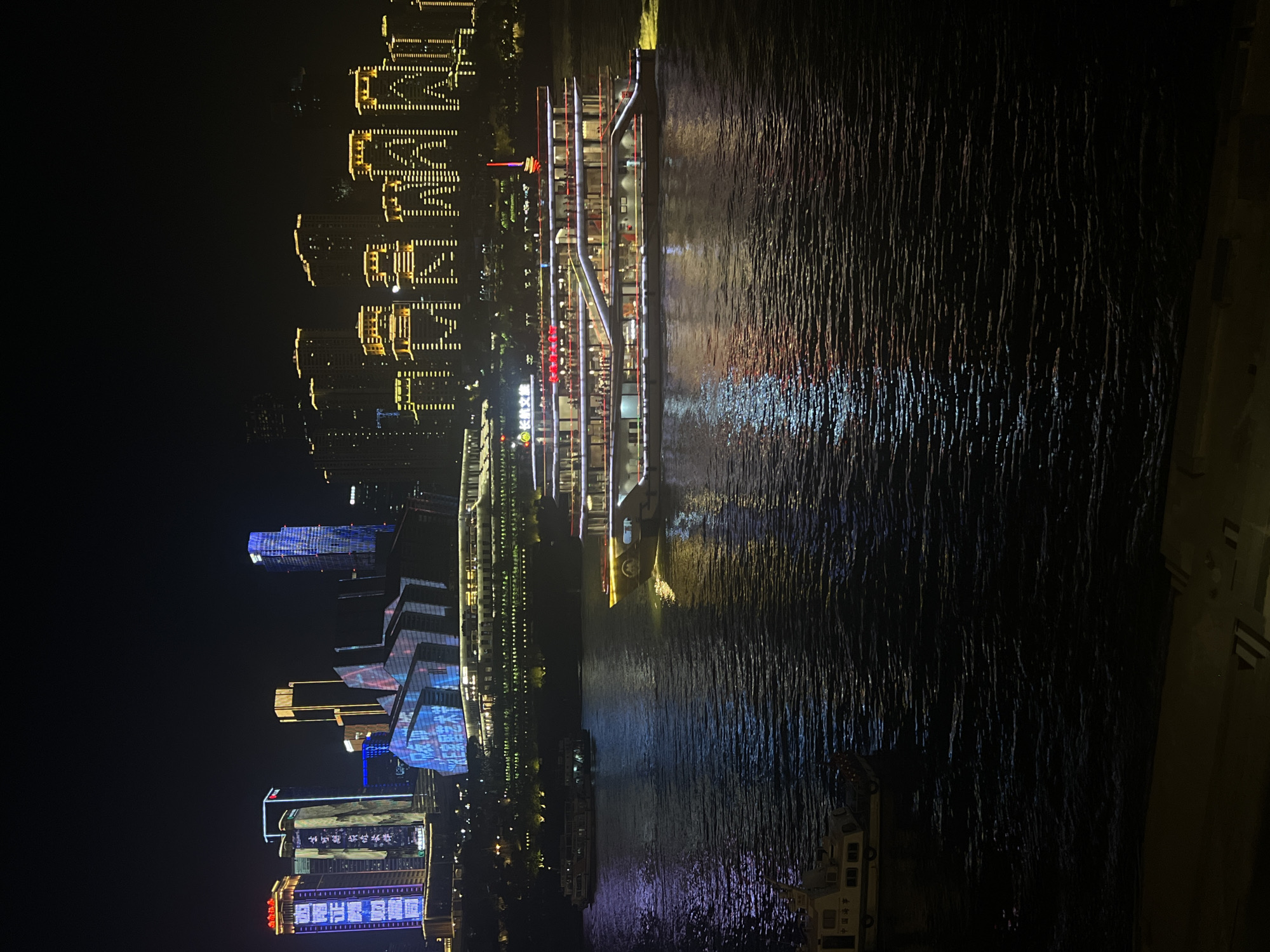 The image shows a nighttime view of the Yuzhong District in Chongqing, China, along the Yangtze River. A large, multi-decked cruise ship is prominently featured in the foreground, illuminated by numerous lights and reflecting on the dark water. The ship has several levels with visible windows and people on board, suggesting it is in operation. Several smaller boats are also present on the river. In the background, the skyline of Yuzhong District is illuminated by a dense collection of skyscrapers and buildings, many of which are lit with colorful lights and projected displays. The overall scene depicts a vibrant urban waterfront at night.