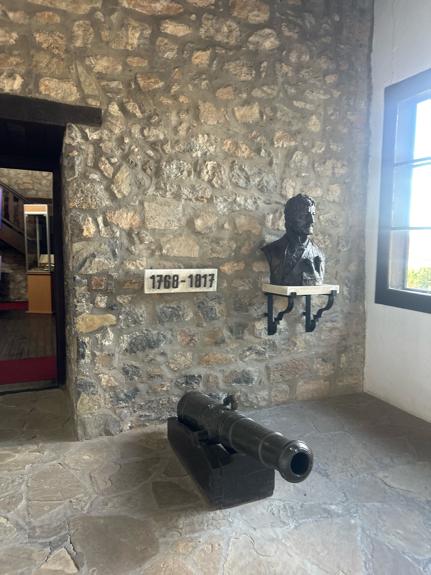 The image depicts an interior space with a stone wall and a stone floor. Mounted on the wall is a plaque with the inscription "1768-1817" and a bust of a man with a mustache and formal attire. To the right of the bust is a window with dark framing, through which sunlight streams. In the foreground, a cannon with a dark barrel rests on a wooden base. To the left of the cannon, an entryway reveals a glimpse of a red carpet and a staircase. This scene is located in the Homeland Museum in Topola Municipality, Serbia, likely within the Oplenac complex, which preserves historical artifacts and exhibits related to Serbian history.