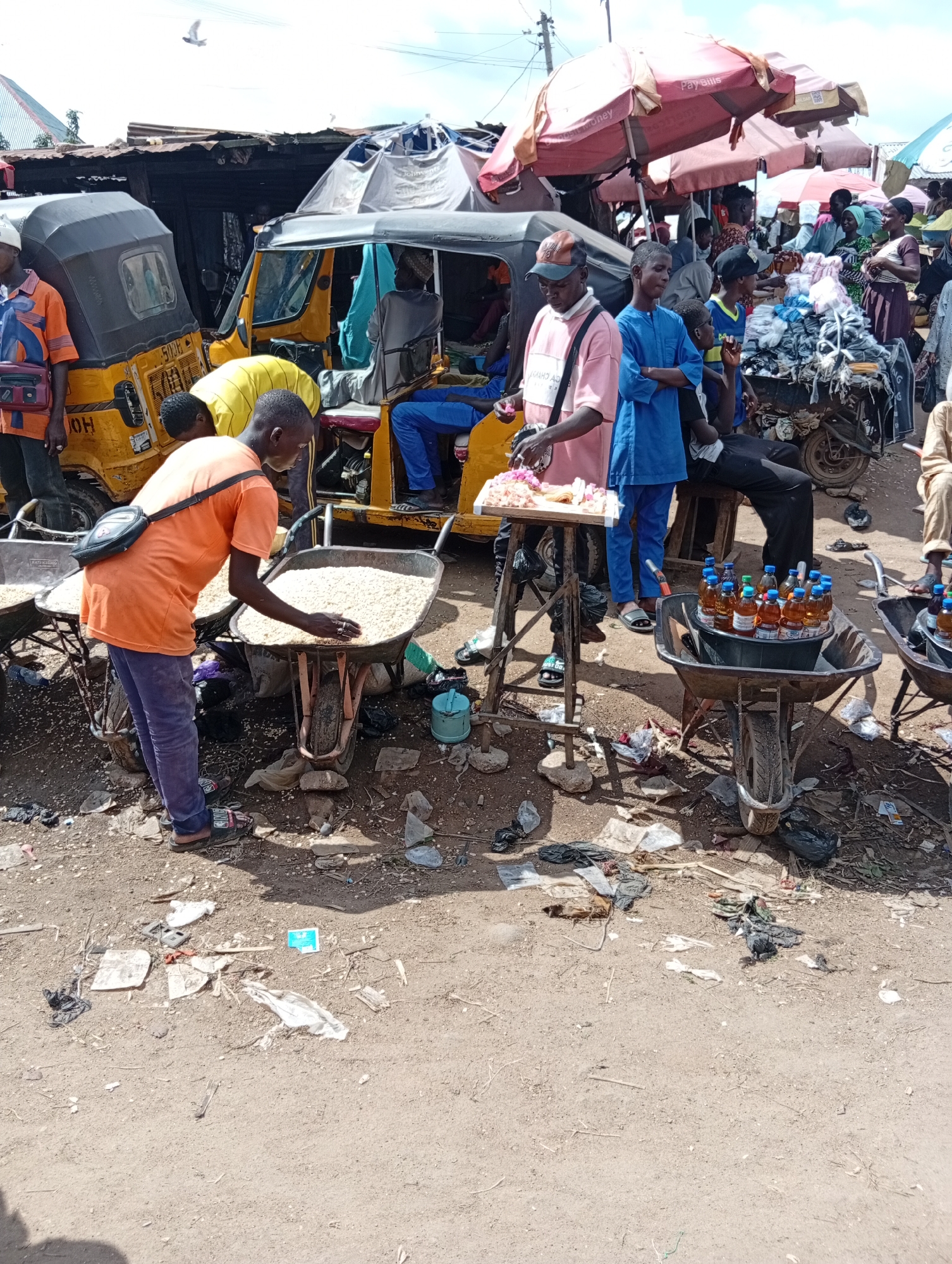This is a wide, outdoor, ground-level shot of a bustling marketplace in Jalingo, Nigeria. The scene is crowded with people, a yellow auto-rickshaw, and various market stalls and goods. The ground is dirt and littered with debris.
In the foreground, a man wearing an orange t-shirt and purple pants is interacting with a wheelbarrow full of light-colored grain. He is reaching into the wheelbarrow with his right hand. To his left, another wheelbarrow, mostly out of frame, also appears to contain grain. Further to the right, a wheelbarrow is filled with an assortment of bottled drinks. A blue bucket holds the bottles, some of which have orange liquid inside.
Behind these foreground elements, a yellow auto-rickshaw with a gray roof is prominent. Several people are either in or around the auto-rickshaw. One man in a pink t-shirt and a cap stands at a wooden table displaying small pink items, possibly food or merchandise. He is interacting with people to his right. A young man in a blue outfit stands with his arms crossed next to him. Another person in a blue shirt is seated behind them.
In the background, more market stalls are visible, with people browsing and interacting. Red and pink umbrellas provide shade. A pile of folded clothing or fabric is stacked on a cart. The overall environment appears to be a busy, informal market setting with a lot of activity. The sky is visible in the upper portion of the image.