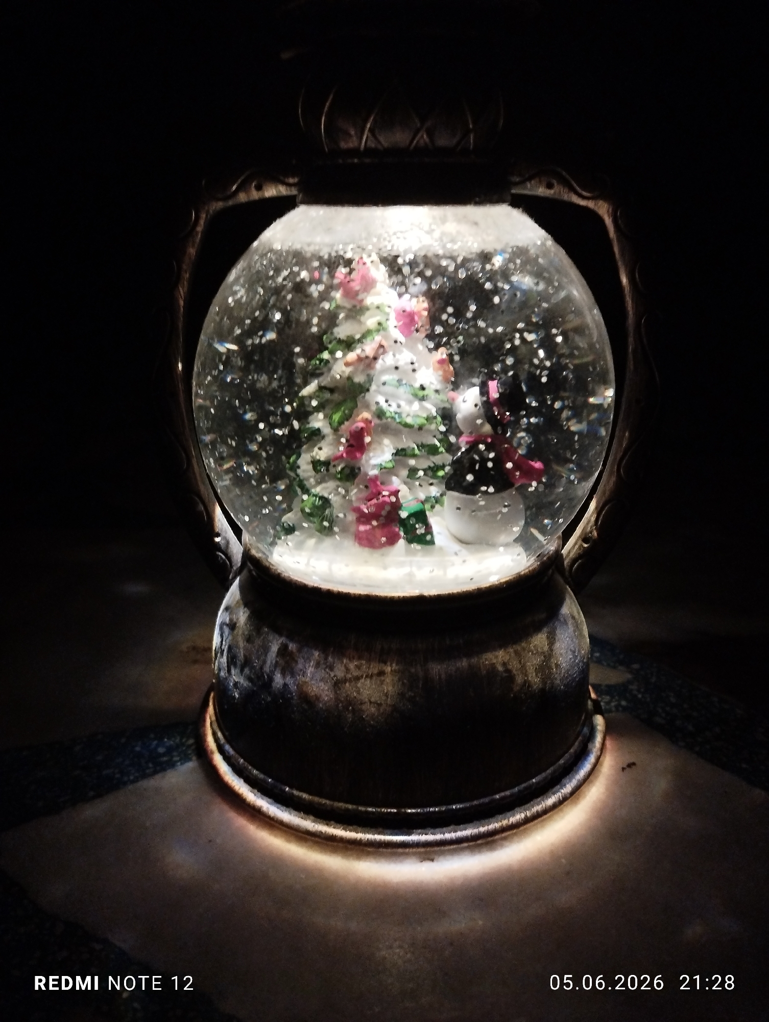 The image displays an internally illuminated snow globe housed within a dark, ornate frame resembling a lantern, positioned in the city of Burgas, Bulgaria. The primary subject, the snow globe, contains a miniature Christmas tree adorned with white "snow," small pink and green decorative elements, and two small figures resembling snowmen, one with a black hat and red scarf. Several miniature gift-like objects (red and green) are visible at the base of the tree. Numerous white and iridescent glitter particles are suspended within the globe, simulating falling snow. The snow globe's internal light source casts a warm glow directly onto the textured, possibly metallic, base of its frame and the surface beneath it. This surface exhibits a patterned, tiled appearance, visible in the lower left and right portions of the frame. The immediate background is dark, providing strong contrast to the illuminated snow globe. Text overlays indicate the capture device as "REDMI NOTE 12" and the date and time as "05.06.2026 21:28". No individuals, actions, or interactions are present in the scene.