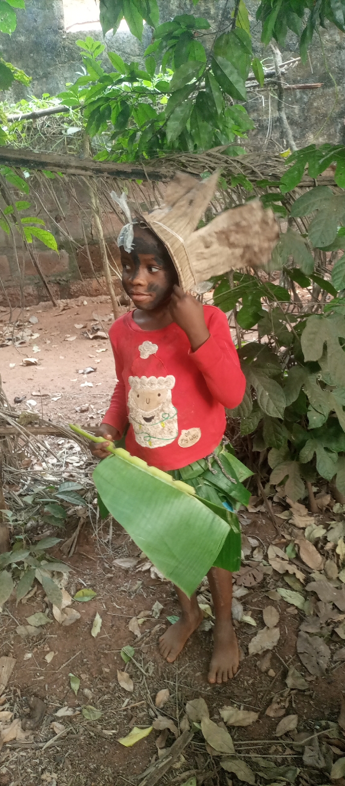 A young person with darkened facial makeup is in a wooded area. They wear a red shirt with a sheep graphic and a leafy skirt. A headdress of straw or dried leaves adorns their head. They hold a large green leaf in their hands, possibly a banana leaf. The ground is dirt-covered and scattered with fallen leaves. Green foliage surrounds the person, suggesting a natural or forest setting. The overall impression is of a costume or role-playing scenario.