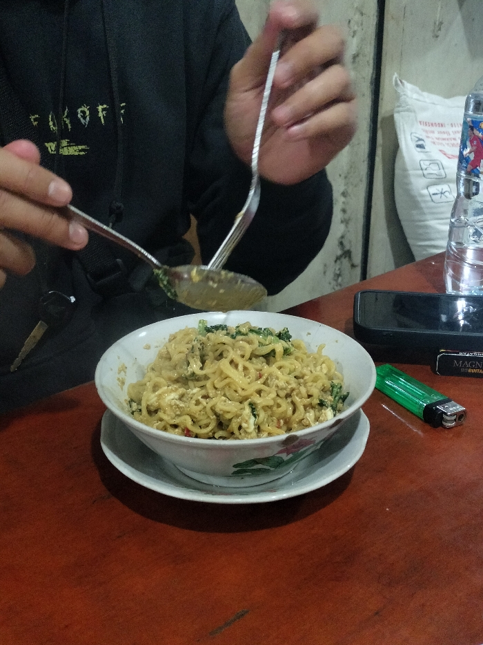 This candid, eye-level shot captures a person eating a bowl of instant noodles in what appears to be a casual indoor setting, likely a small eatery or *warung* in Dramaga, Indonesia. The dominant subject is a person, visible from the mid-torso upwards, wearing a black hoodie with partially visible white-green text that reads "F*CKOFF". Their hands are engaged in the act of eating, using a spoon and a fork to scoop noodles from a white ceramic bowl.
The main dish is a generous serving of instant noodles, seemingly fried or dry-style (*mie goreng*), mixed with bits of green leafy vegetables (possibly bok choy or similar) and scrambled egg. The bowl has a subtle floral design around its rim and rests on a matching white saucer.
On the wooden table, alongside the noodle bowl, are several personal items. To the right, a black smartphone rests on its side, next to a clear plastic water bottle with a blue and red label. Further to the right, a black pack of "MAGNUM" "BINTANG" cigarettes and a bright green lighter are visible. Car keys hang from the left side of the table, near the person's left hand.
The background is simple, with a light-colored, somewhat textured wall on the right side and a large white sack, possibly containing rice or other dry goods, leaning against it. The lighting is soft and ambient, suggesting it might be evening or night. The overall scene conveys a common, everyday meal experience in Indonesia.