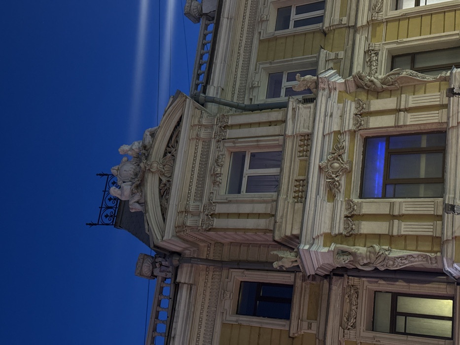 The image depicts the ornate facade of the 'House with Atlantes' located at Solyanka Street, 1/2, building 2, Moscow, Russia. The scene is set during evening or nighttime, under a deep indigo-blue sky with faint, elongated white light streaks.
The multi-story building features a light yellow/beige exterior adorned with extensive white classical revival architectural details and ornamentation. At the roofline, a prominent central structure is crowned by three large, light-colored sculptural figures: a central shield or cartouche flanked by two reclining classical human figures. Above this, a dark, decorative wrought-iron railing is visible.
Below the roofline, the facade displays multiple rectangular windows with elaborate white architraves, cornices, and pilasters. Interior lighting is visible through some windows, with a distinct blue hue emanating from a lower-level window and yellowish light from others. Architectural details include a caryatid (a sculpted female figure) supporting a structural element on the building's prominent right corner, and a smaller winged figure positioned on a ledge above a window. A dark vertical downspout runs along the left corner of the building. The facade is illuminated, accentuating its intricate sculptural and architectural elements. No individuals or discernible activities are present within the frame.