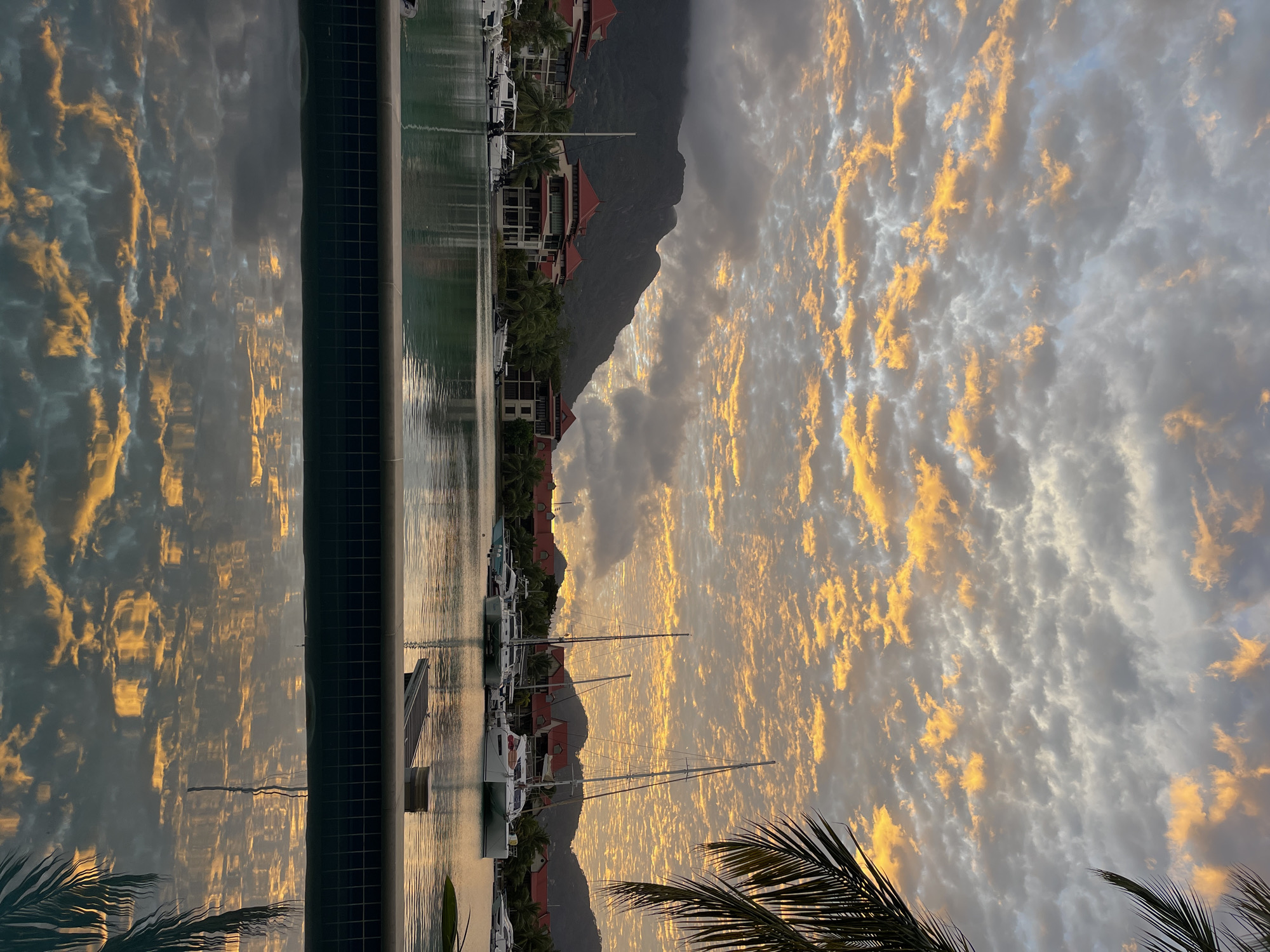 The image depicts a serene harbor scene in Victoria, Seychelles, at what appears to be dusk. The sky is filled with dramatic clouds, illuminated by the warm hues of sunset, casting an orange glow on the scattered white clouds against a backdrop of gray. In the mid-ground, several sailboats and yachts are moored in the tranquil water, their masts reaching towards the colorful sky. On the shore, a row of buildings with distinctive red-tiled roofs, characteristic of tropical architecture, are visible, interspersed with lush green palm trees and other vegetation. Behind the buildings, a range of dark, forested mountains forms the distant horizon, partially obscured by some clouds. The foreground features the edge of a swimming pool, its surface reflecting the vibrant sky and the scene beyond, creating a mirror image of the harbor and the sunset. A palm frond is visible on the right side of the frame, adding to the tropical ambiance. There are no people visible in the frame. The overall atmosphere is one of peacefulness and natural beauty.