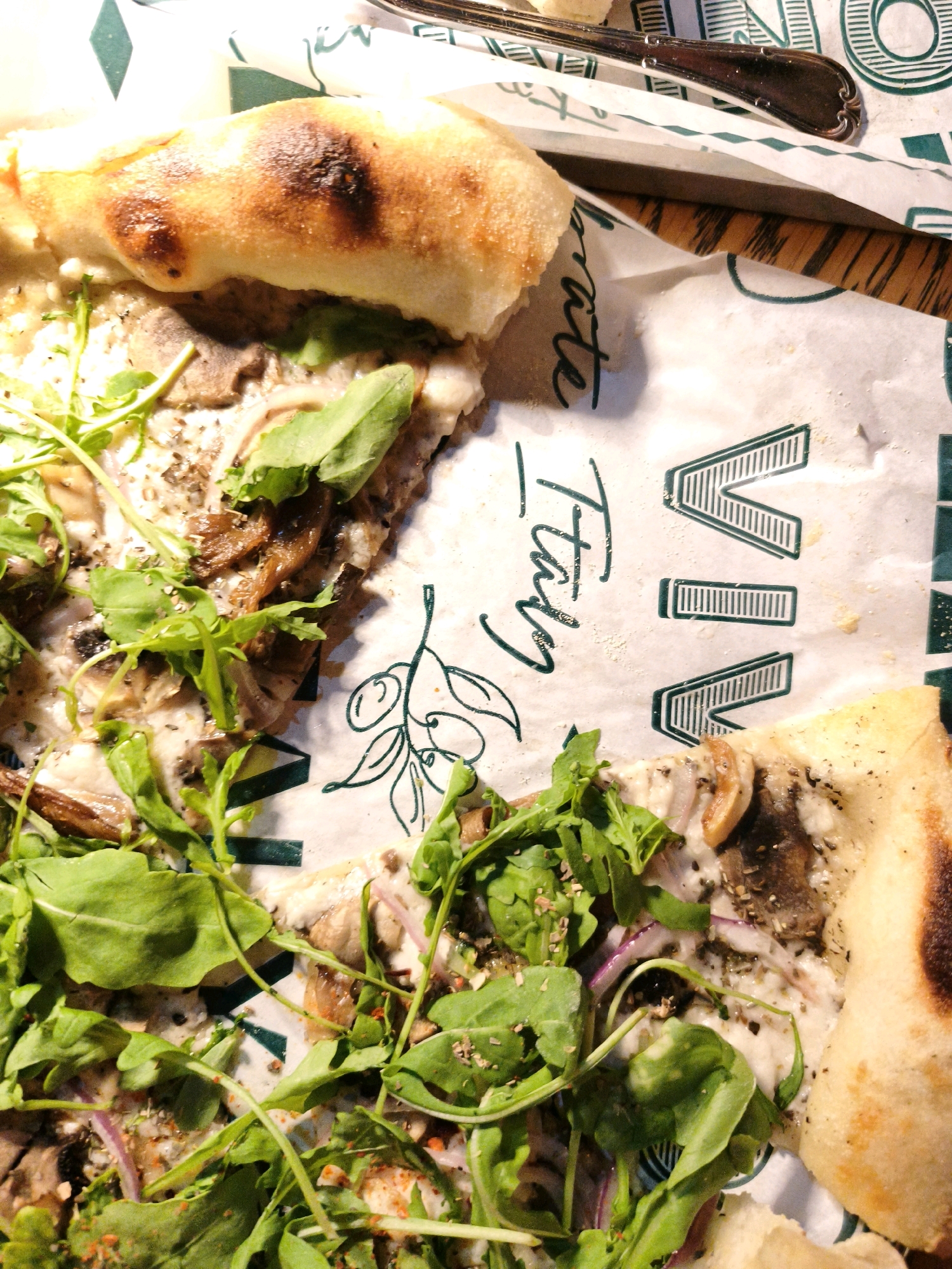 The image shows a close-up view of a pizza with arugula and mushrooms on parchment paper. The pizza has a slightly burnt crust and appears to be served on a table with a fork nearby. The background is blurred, but suggests an indoor setting. There are no people visible in the image. The text on the parchment paper, "VIVA Italia," indicates a possible connection to Italian cuisine. The image does not provide any information about the location of the city of Be'er Sheva, Israel.