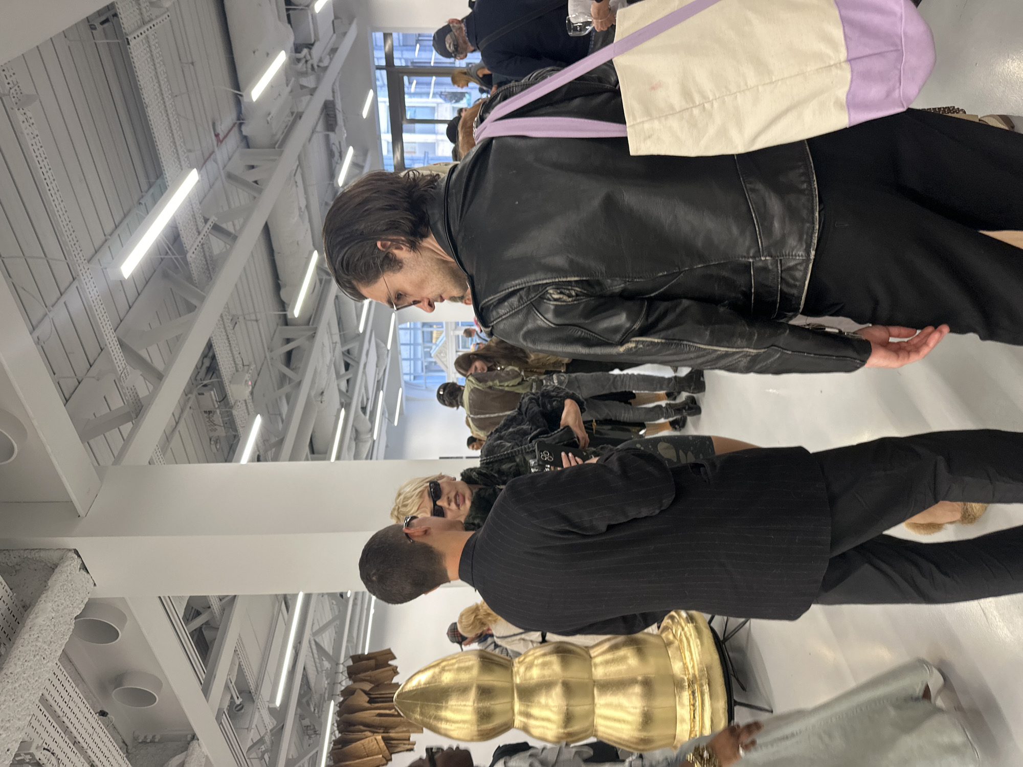 Set in London, United Kingdom, this image captures a bustling indoor event, possibly an art exhibition or social gathering. In the foreground, a man with dark hair and glasses, wearing a black leather jacket and black trousers, is seen from the side, looking downward. He carries a white and light purple tote bag slung over his shoulder. Next to him, with his back to the viewer, another man in a dark pinstripe suit and black trousers stands, seemingly engaged with a blonde-haired woman in sunglasses, who is wearing a dark, fur-like jacket and holding a mobile phone.
To the left, a large, golden, segmented sculpture occupies the midground. Behind these figures, several other people are visible, some blurred, suggesting a crowded environment. One individual in the background wears a cap and holds a glass. The setting features a high white ceiling with exposed structural beams, white ventilation ducts, and numerous rectangular fluorescent light fixtures, giving the space an industrial-chic aesthetic. Natural light streams in from a large window visible in the background, indicating it is daytime. On a surface to the left, stacks of brown paper bags are neatly arranged. The overall atmosphere appears lively and modern. No discernible text is visible within the frame.