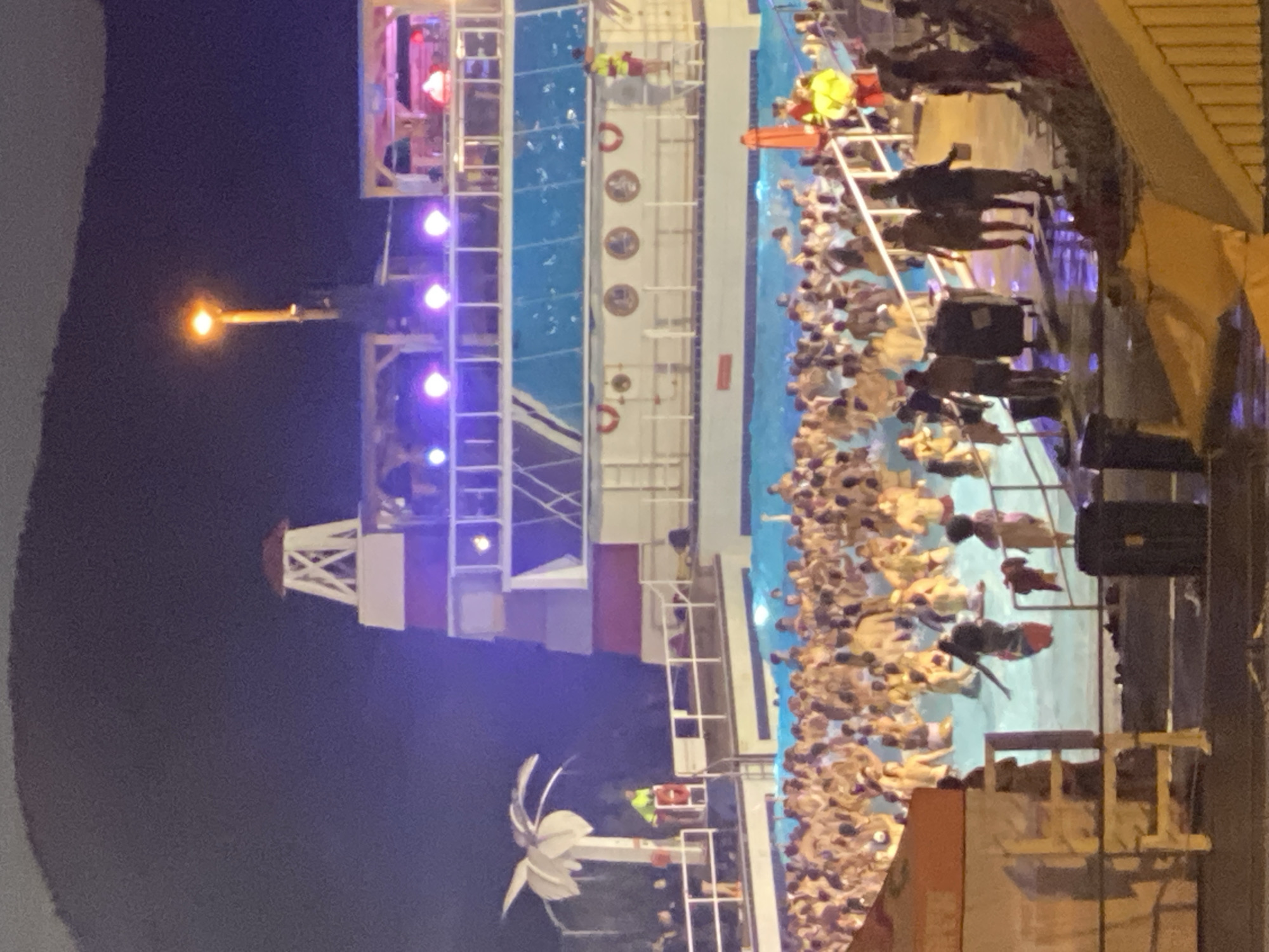 The image depicts a crowded outdoor swimming pool at night, likely at the Village Vacances Valcartier in Saint-Gabriel-de-Valcartier, Canada, given the presence of a distinctive multi-story structure resembling a themed water park building, possibly a replica ship or lighthouse, illuminated by purple and white lights. A large number of people, predominantly young adults, are in the water, with many standing and interacting. Some individuals appear to be swimming or wading. The pool area is surrounded by railings and a deck where other people are standing or walking. Several lifeguards in bright yellow vests are visible, some observing the pool from elevated positions. In the background, a dark, undulating hillside or mountain range is visible against the night sky, illuminated by a bright street lamp. The overall atmosphere suggests a lively nighttime event or party at a water park.