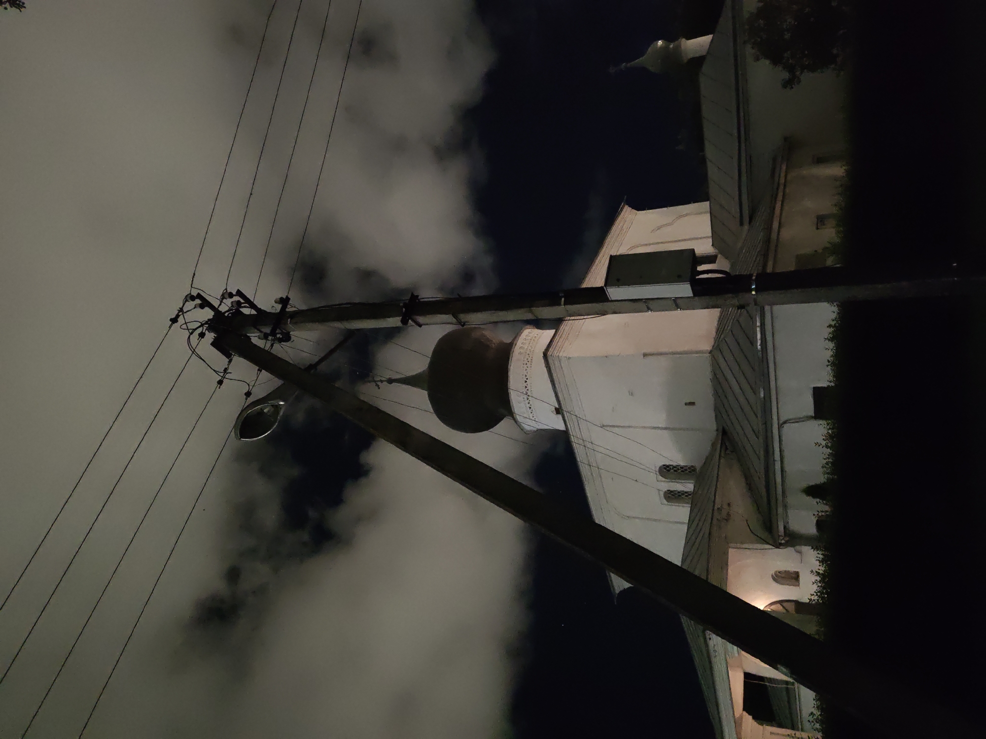 The image shows a night view of the Church of St. Sergius of Radonezh (Tserkov' Sergiy Radonezhskogo) in Pskov, Russia. A wooden utility pole with multiple wires stretches across the foreground, leaning diagonally from the lower left to the upper right. Behind the pole, the white facade of the church is visible, with a dark, bulbous dome topping its central structure. The church has a sloping, gray metal roof. The sky is dark, with patches of illuminated clouds. A smaller, green-domed structure, likely another part of the church complex, is visible to the right. The lower portion of the image is in darkness, obscuring the ground level. No people are visible in the scene.