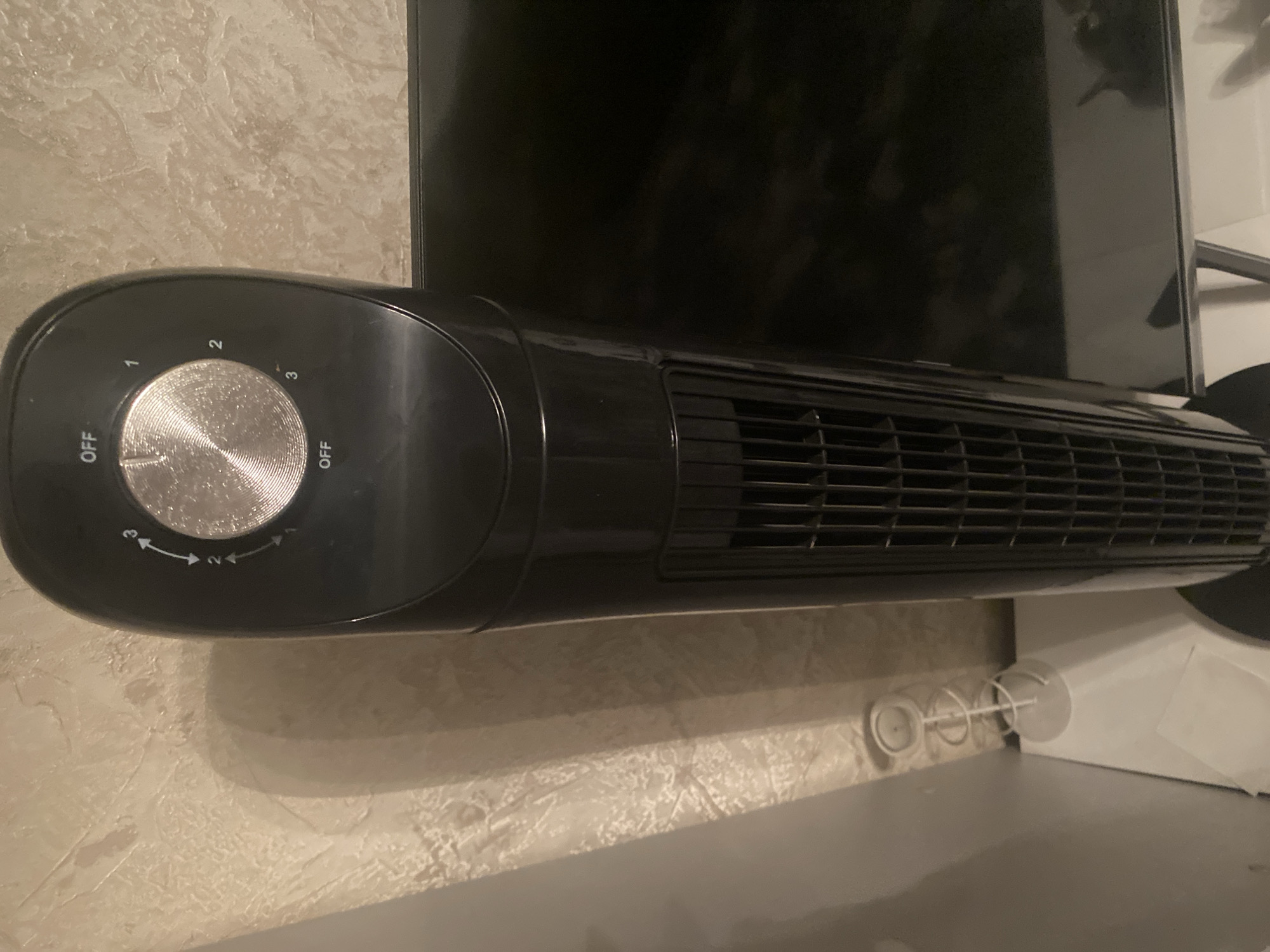 The image shows a black tower fan positioned vertically. The top of the fan features a control dial with settings labeled "OFF," "1," "2," and "3." The dial is currently set between "OFF" and "1." The fan is placed in front of a textured, light-colored wall. To the left of the fan, a white, spiral-shaped object, possibly a decorative item or candle holder, is visible on a white surface. A dark, reflective surface, likely a television screen, is to the right of the fan. The scene appears to be indoors, with soft lighting casting shadows. The specific location within Chernogorsk, Russia, is not determinable from the provided image.