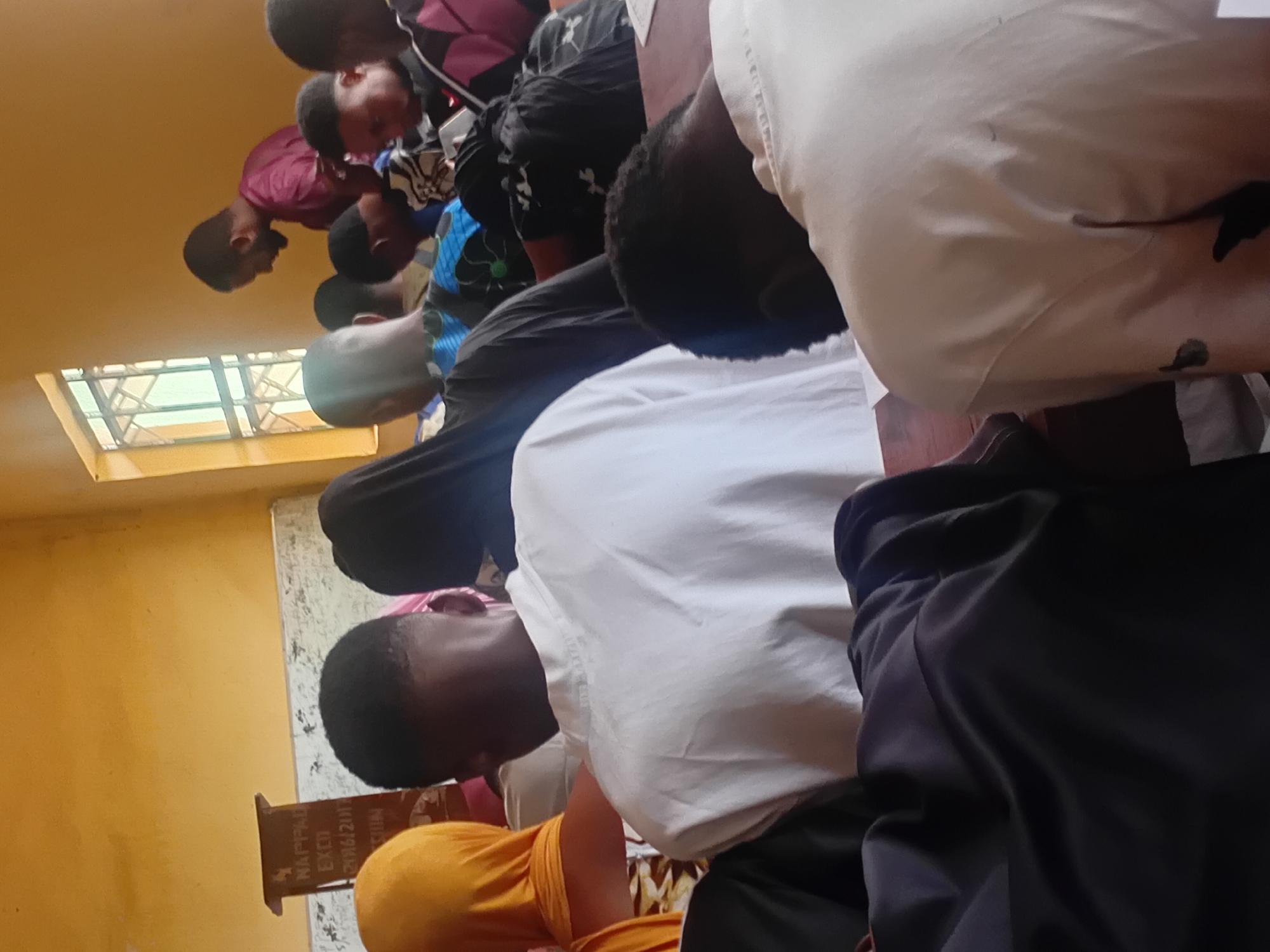 The image depicts an indoor scene, possibly a classroom or meeting hall, with several individuals seated. The room has yellow walls and a window with barred panes. In the foreground, the backs of people wearing predominantly white and dark clothing are visible, suggesting they are facing away from the camera. A lectern is visible on the left side of the image with some writing on it. On the right, a man in a maroon shirt is standing and looking down. The overall atmosphere suggests a gathering or a lecture. This scene is located in Ogbogbo, Nigeria.