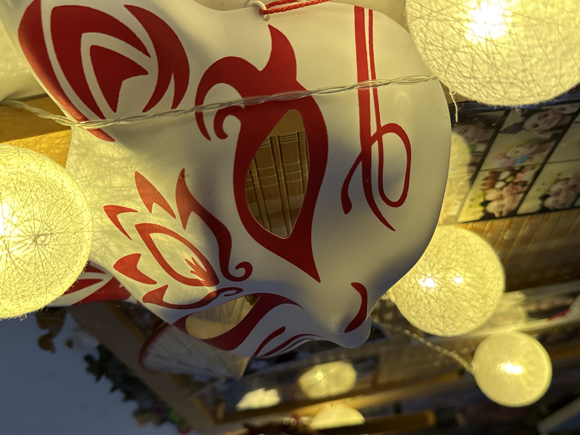 The image is an upside-down, close-up shot of a white Japanese Kitsune mask with red markings. The mask features intricate red designs around the eyes and on the cheeks, typical of traditional Kitsune masks, often associated with foxes in Japanese folklore. The mask has eye openings that reveal a glimpse of what appears to be a bamboo-like texture inside. A thin string of fairy lights, with several spherical, woven lights, is draped across the mask, casting a warm glow.
In the background, some out-of-focus items are visible. To the right of the mask, there is a display of what looks like framed pictures or cards, with blurry images of faces, possibly cartoon characters or people. The overall setting appears to be indoors, likely a shop or a decorated space, given the display and the ambient lighting. The reflections on a glossy surface below suggest the photo might have been taken on a table or shelf. The lighting is warm and yellow, emanating from the fairy lights, creating a cozy or festive atmosphere. There is no visible text in the image. The orientation of the image, being upside down, adds a unique perspective to the scene. The subjects are the mask and the decorative lights, with the background elements providing context to the setting. No specific activity or event is clearly depicted, but the presence of decorative lights and the mask suggests a possible cultural event, a festival, or simply a decorative display.