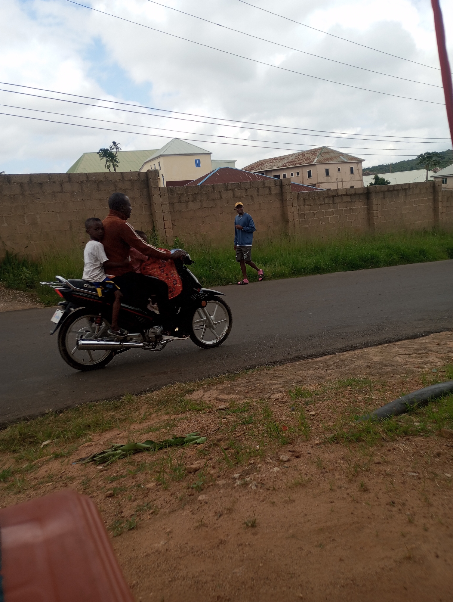 The image depicts a street scene in Jos, Nigeria. A man and a child are riding on a black motorcycle. The man, wearing a brown long-sleeved shirt and dark trousers, is driving. The child, wearing a white t-shirt and blue shorts, is sitting behind him. The child is looking towards the camera. The motorcycle is moving on an asphalt road. To the left of the road, there is a brown brick wall and a patch of green grass. In the background, there are buildings with corrugated iron roofs and some trees. Another person, wearing a blue hoodie and a yellow cap, is walking on the right side of the road, facing away from the camera. The sky is cloudy. In the foreground, there is a reddish-brown object, possibly a piece of furniture, and some loose soil with scattered green vegetation.
