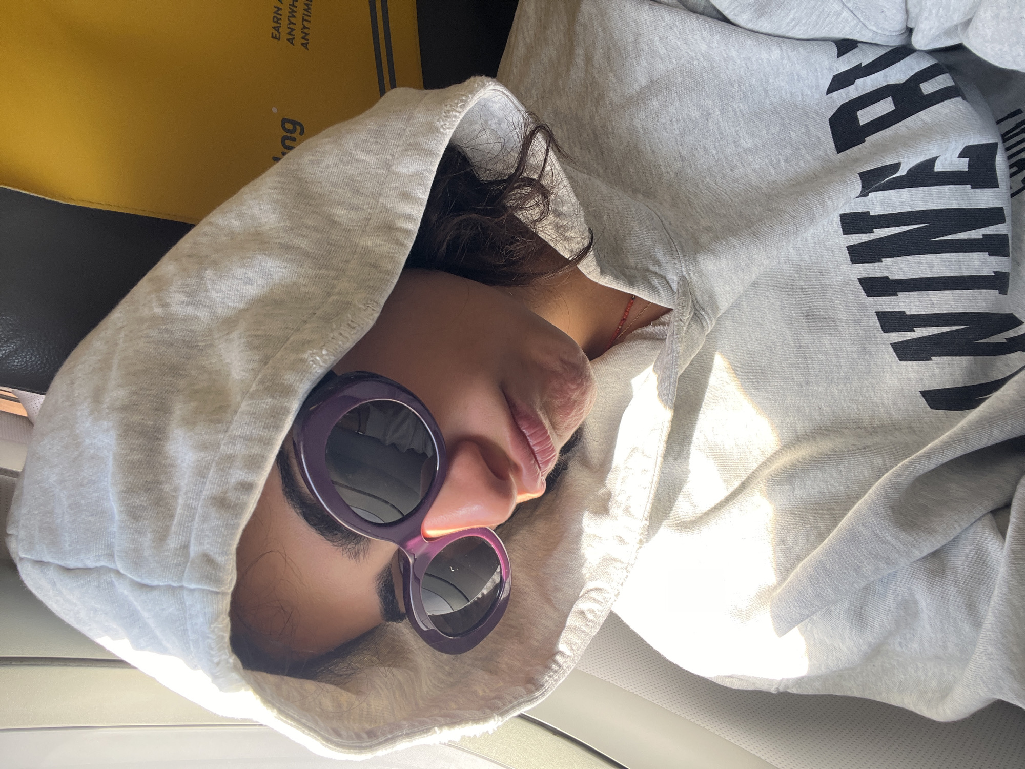 A young person wearing a light gray hooded sweatshirt and oversized purple sunglasses is looking down and to the side. The person's head is partially obscured by the hood. Sunlight is coming in from the left, casting shadows. In the background, a yellow surface with some text and a black object are visible. The scene appears to be inside a vehicle, possibly a bus or train, given the presence of a seat and window. The text on the yellow surface is partially obscured but includes "EARN A" and "ANYWH" suggesting an advertisement. The overall impression is of someone trying to rest or relax. The location is not determinable from the visual information provided.