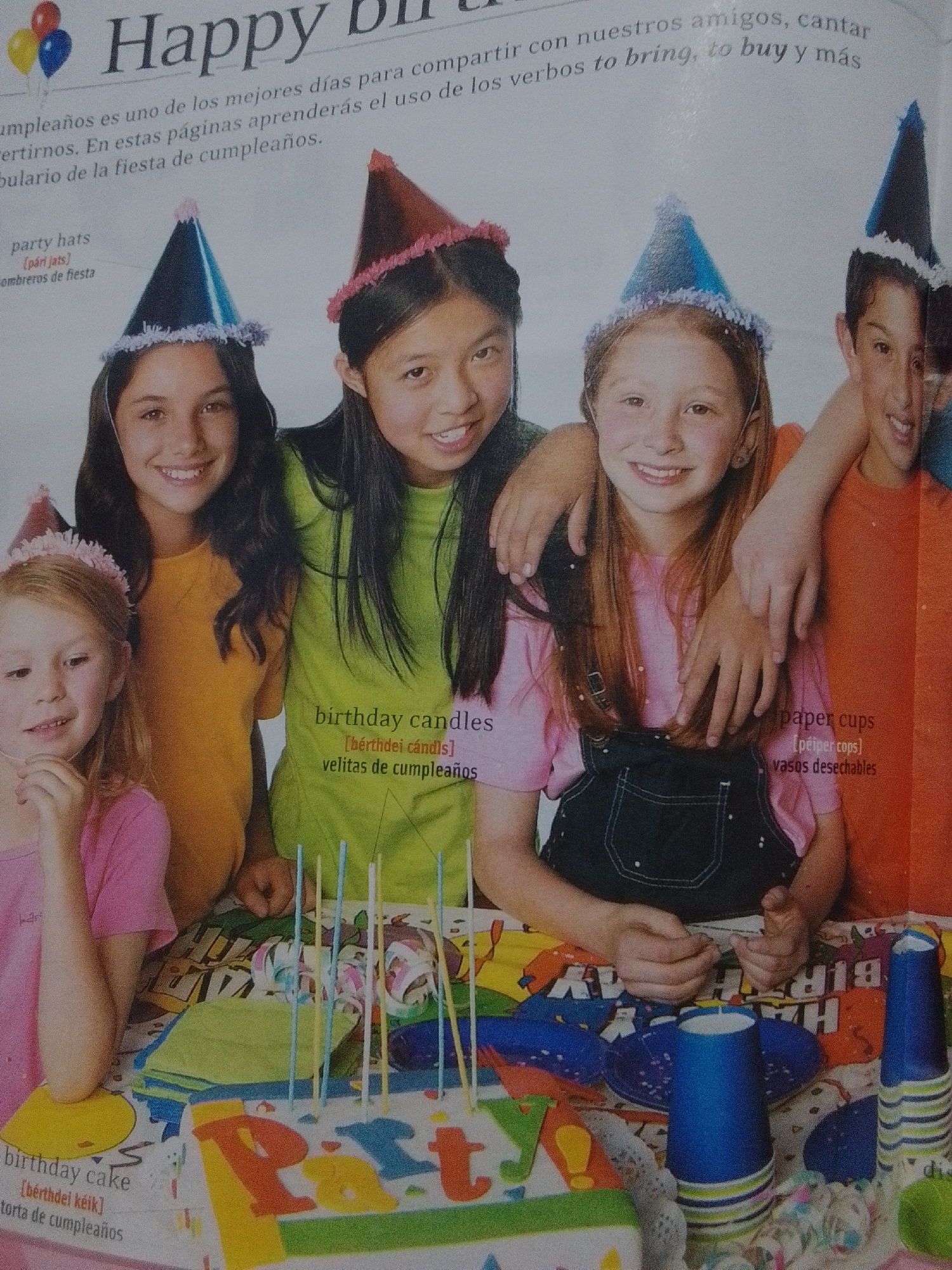 The image depicts a group of four children, three girls and one boy, wearing party hats and posing for a photo. They are gathered around a birthday cake on a table covered with a colorful, printed tablecloth. The children appear to be celebrating a birthday party. In the background, text is visible, suggesting this is from a magazine or educational material, likely related to learning English. The scene is indoors, with artificial lighting. No information about the location being the city of La Tagua, Venezuela, is present in the image.