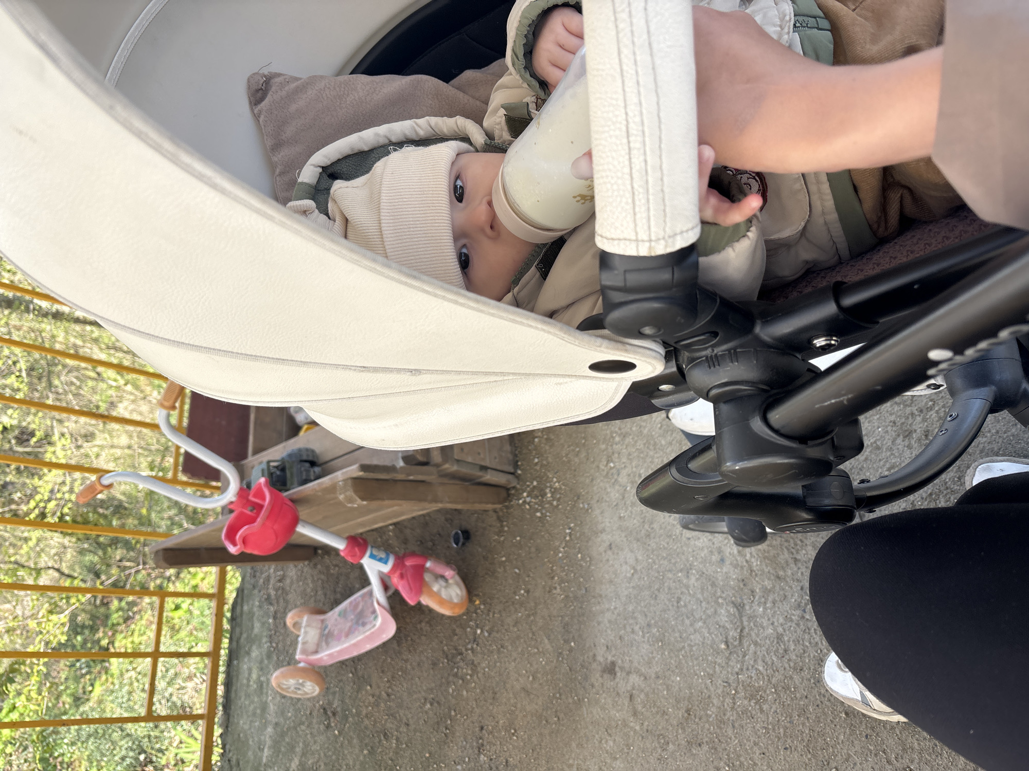 A baby in a tan jacket and hat is being bottle-fed in a white baby stroller. The bottle is white with golden markings. A woman's arm is visible holding the bottle. The stroller is on a paved surface. Adjacent to the stroller, a pink and white child's scooter sits near a yellow metal railing, suggesting a balcony or elevated walkway. The scene appears to be outdoors, with hints of greenery visible behind the railing. There's wooden platform behind the scooter. Without additional context, Sochi, Russia cannot be confirmed.