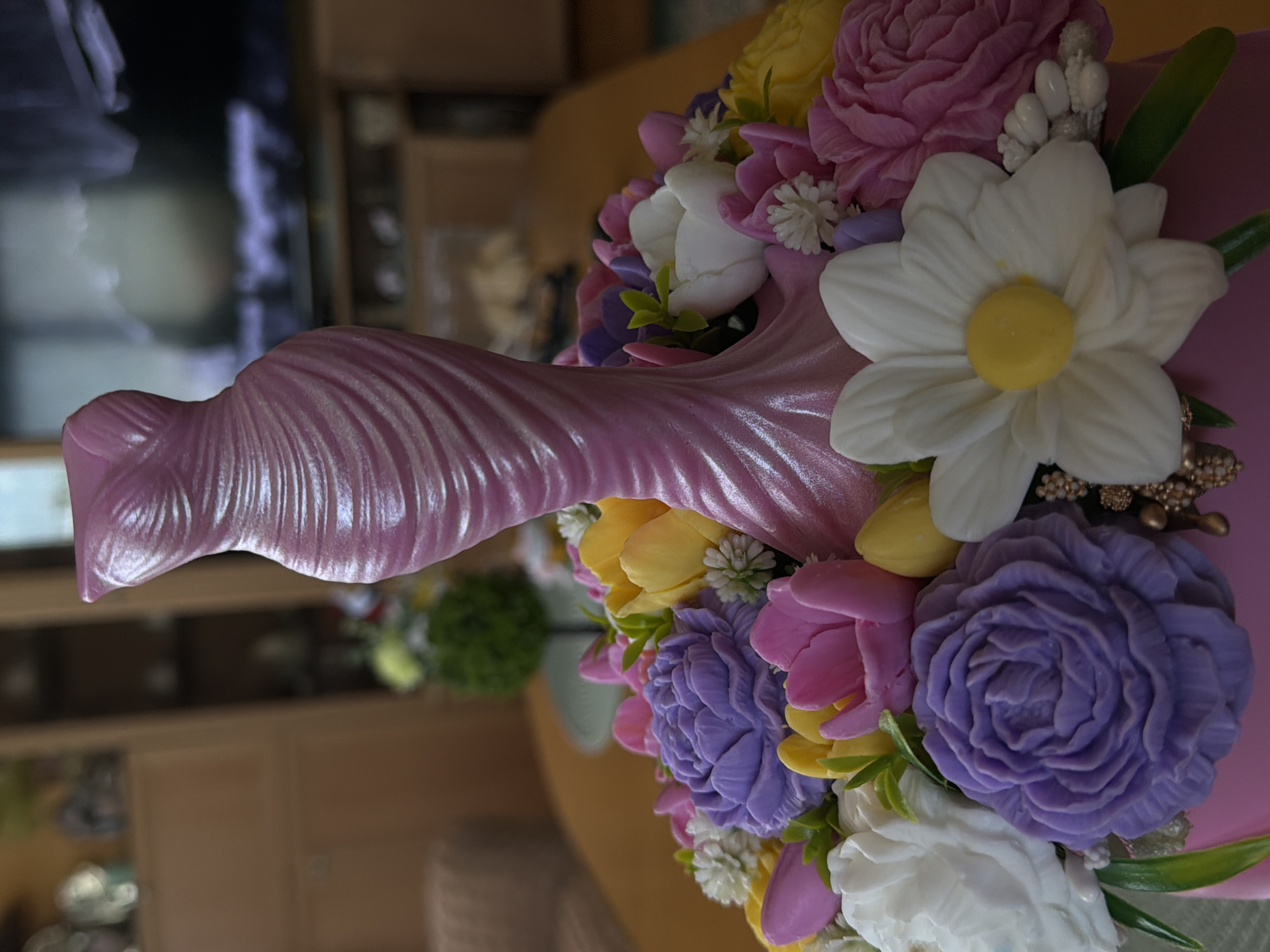 A close-up, eye-level, indoor shot showcases a decorative pink cake shaped like a flowing dress, adorned with an assortment of intricately crafted soap flowers in vibrant shades of purple, pink, yellow, and white. The soap flowers are arranged around the base of the dress-shaped cake. The background is blurred, suggesting an indoor setting with furniture and possibly a television. The lighting is soft, highlighting the pearlescent sheen of the dress cake and the delicate details of the soap flowers. There is no indication of the city of Lyubimets, Bulgaria, in this image.