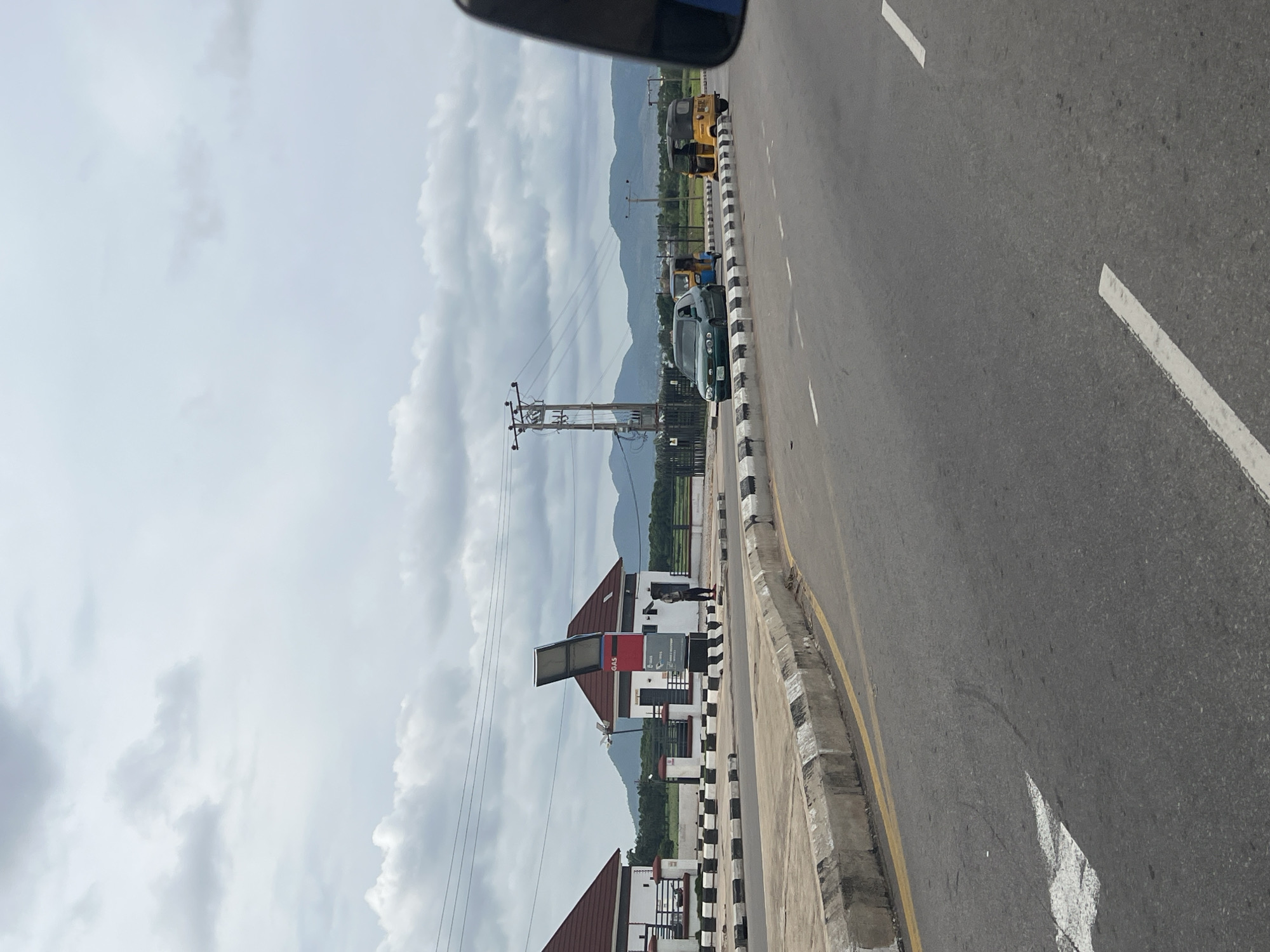 The image depicts a roadside scene in Jalingo, Nigeria, under a cloudy sky. In the foreground, a grey asphalt road with white lane markings and a yellow dividing line is visible. A concrete curb with black and white hazard stripes runs along the side of the road.
On the left, a modern-looking building complex with white walls and red-tiled roofs is situated behind a black and white striped barrier. Adjacent to this complex, a fuel station sign with "GAS" clearly displayed is prominent. A person stands near the fuel station.
In the mid-ground, several vehicles are present on the road. A dark green sedan is moving past the fuel station. Behind it, two yellow auto-rickshaws (Keke Napep) are also on the road, with one slightly ahead of the other. A power pole with electrical lines stands further back.
The background shows a line of distant mountains under the overcast sky. The overall environment appears to be a developed area along a major road.