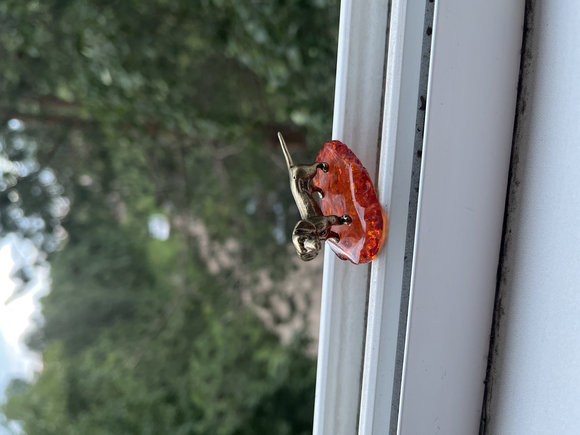 A close-up, eye-level, outdoor shot shows a miniature brass dachshund figurine standing on a translucent amber-colored base. The base is irregular in shape and appears to be made of resin, with small, dark inclusions visible within it. The dachshund is positioned facing left, with its tail extended straight behind it. The figurine and its base are resting on a white windowsill. Beyond the windowsill, a blurred background of lush green trees and foliage is visible, suggesting the location is outdoors with natural scenery. The sky is partially visible at the top of the frame, appearing bright and possibly overcast. The lighting suggests it is daytime.