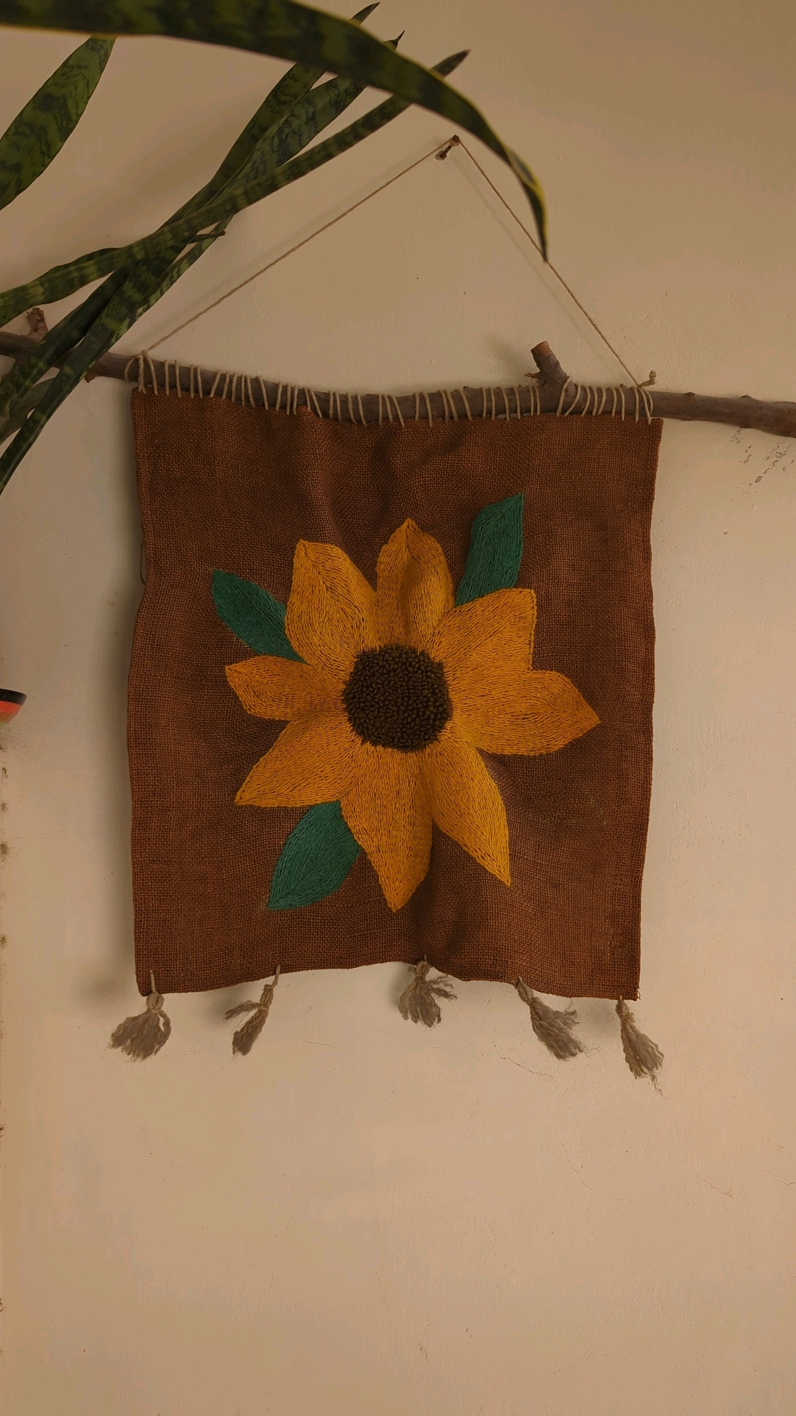 A square, brown, burlap-like tapestry with an embroidered yellow sunflower and green leaves hangs from a thin branch attached to a white wall. The tapestry is suspended by a thin, beige string tied to the branch, which is itself attached to the wall with a small nail. The top edge of the tapestry is finished with loops of beige string. Three beige tassels hang from the bottom corners of the tapestry. A portion of a long, green, leafy plant is visible in the upper left corner of the image, its leaves drooping over the tapestry. The wall appears to be textured and slightly cracked. This scene is located in Wah Model Town Phase 1, Pakistan.