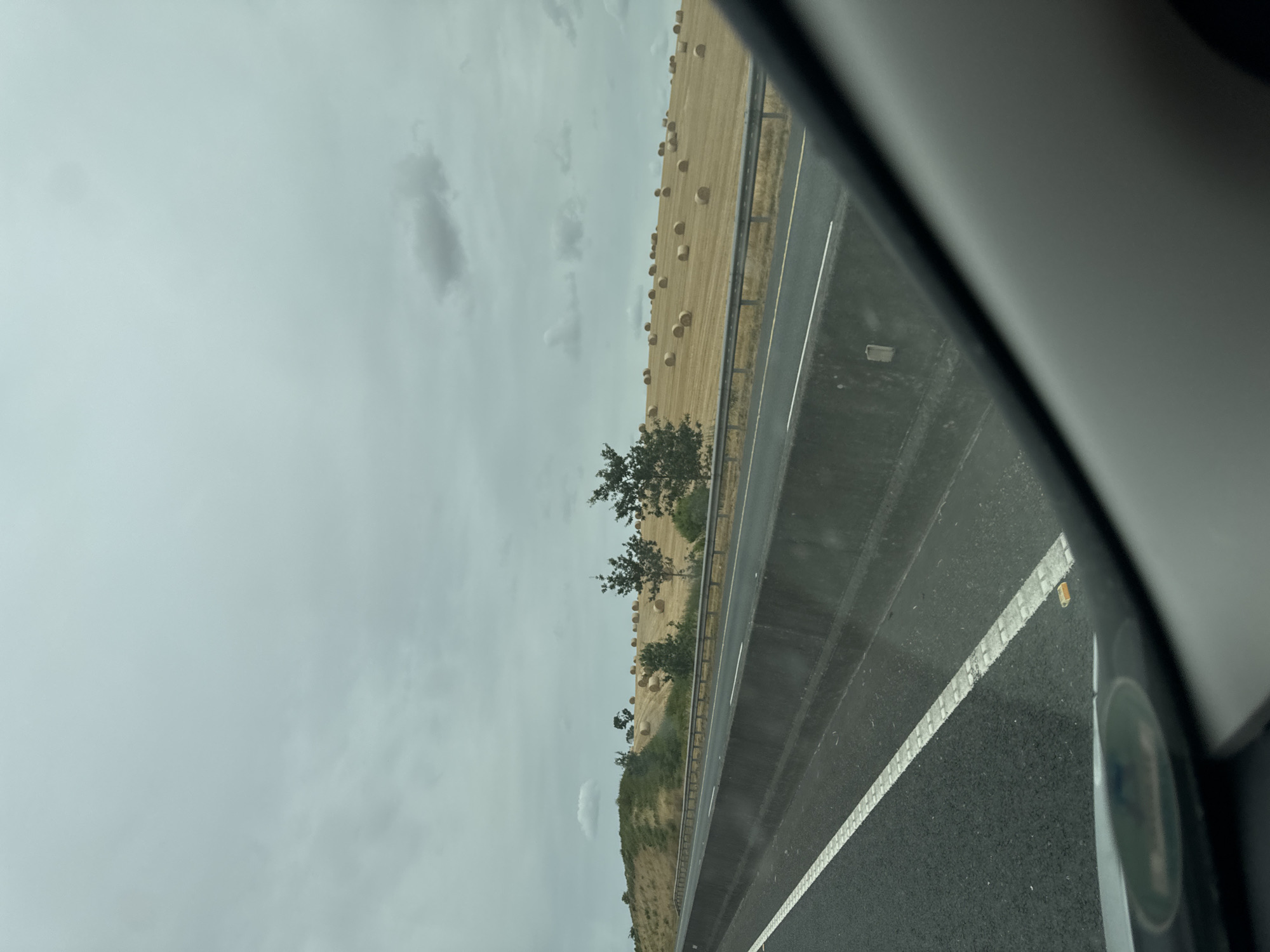 The image captures a view from a vehicle driving on a highway in Ireland, looking towards a rural landscape. The highway itself is paved with asphalt and marked with white lines. A concrete barrier separates the lanes of traffic. Beyond the barrier, a grassy embankment slopes upwards. The landscape is dominated by a field of harvested crops, with numerous round hay bales scattered across it. Several trees are visible, some scattered among the hay bales and others on the higher ground. The sky is overcast with clouds. The image appears to be taken during daylight hours.