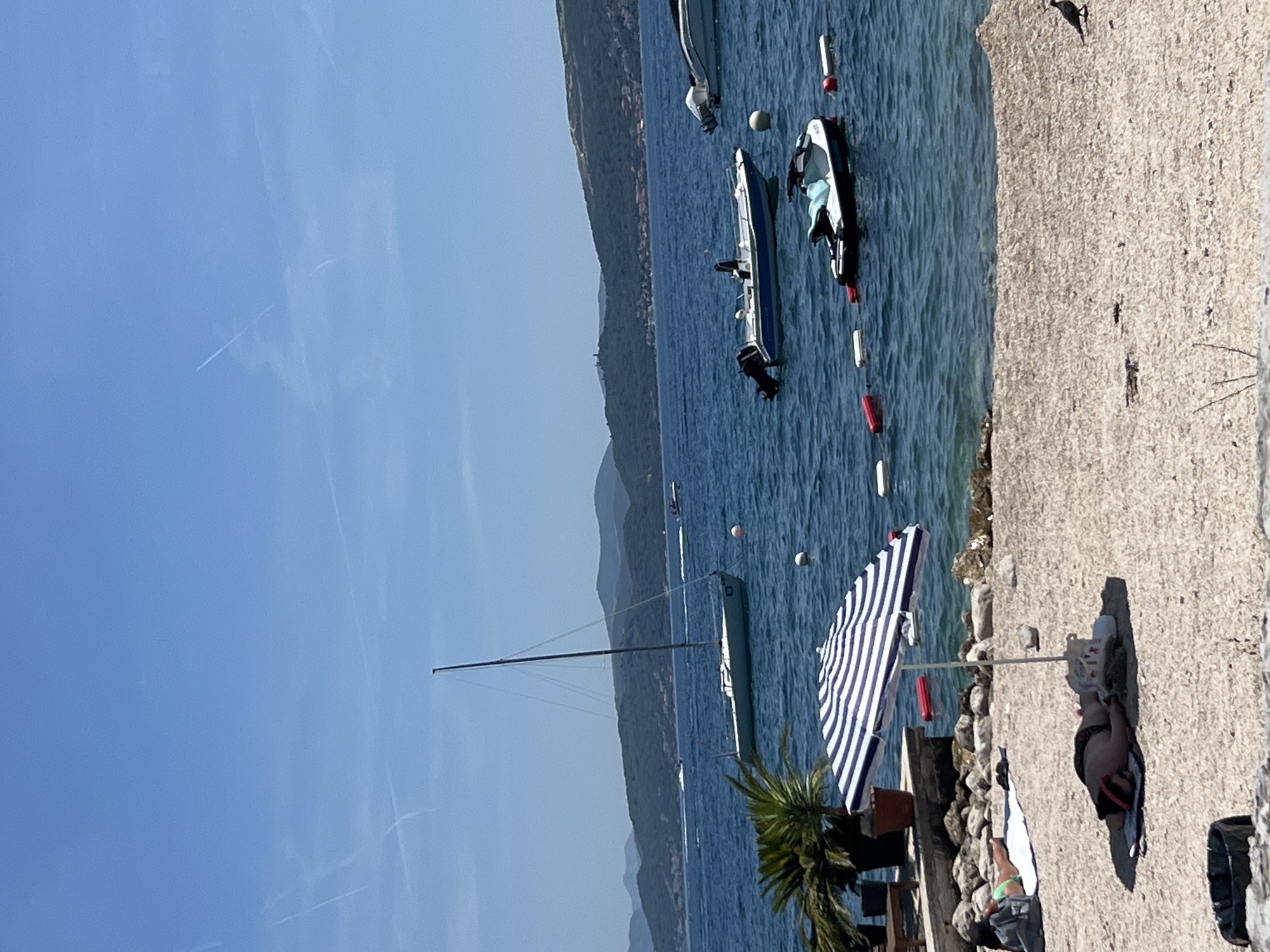 The image depicts a sunny day scene at a beach in Baošići, Montenegro. In the foreground, a sandy beach is visible with two individuals sunbathing. One person is lying on their back under a blue and white striped umbrella, while another person is lying on their stomach on a towel further back.
The water in the background is a deep blue, with several boats visible. A sailboat with a tall mast is anchored offshore, and several smaller motorboats and a jet ski are also present. A line of buoys separates the swimming area from the boats. In the distance, rolling green hills covered with vegetation and scattered buildings form the coastline. The sky is clear blue with a few faint contrails from airplanes. The overall atmosphere is one of leisure and relaxation on a summer day.