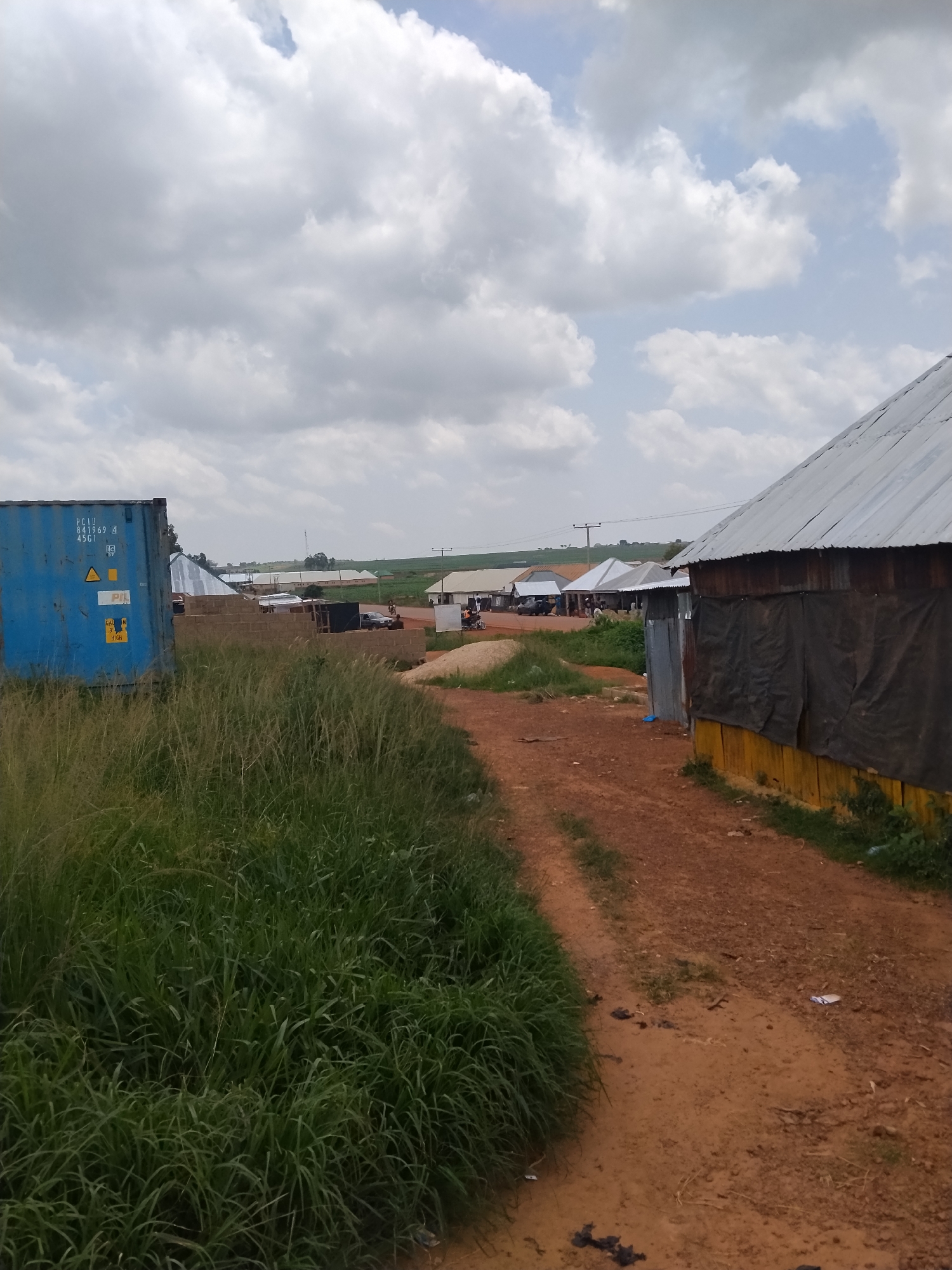 The image depicts an outdoor scene in what appears to be the city of Durumi, Nigeria. The foreground is dominated by a dirt path winding through tall green grass on the left. To the right, a structure with a corrugated metal roof and a yellow base covered by black tarp is partially visible. Further into the scene, more buildings with metal roofs line the path, suggesting a built-up area. In the mid-ground, there's a large blue shipping container with some markings on its side. Several vehicles, including what looks like a car and a motorcycle, are parked along the path, and a few individuals can be seen near the structures. The background shows more buildings and a field under a cloudy sky.