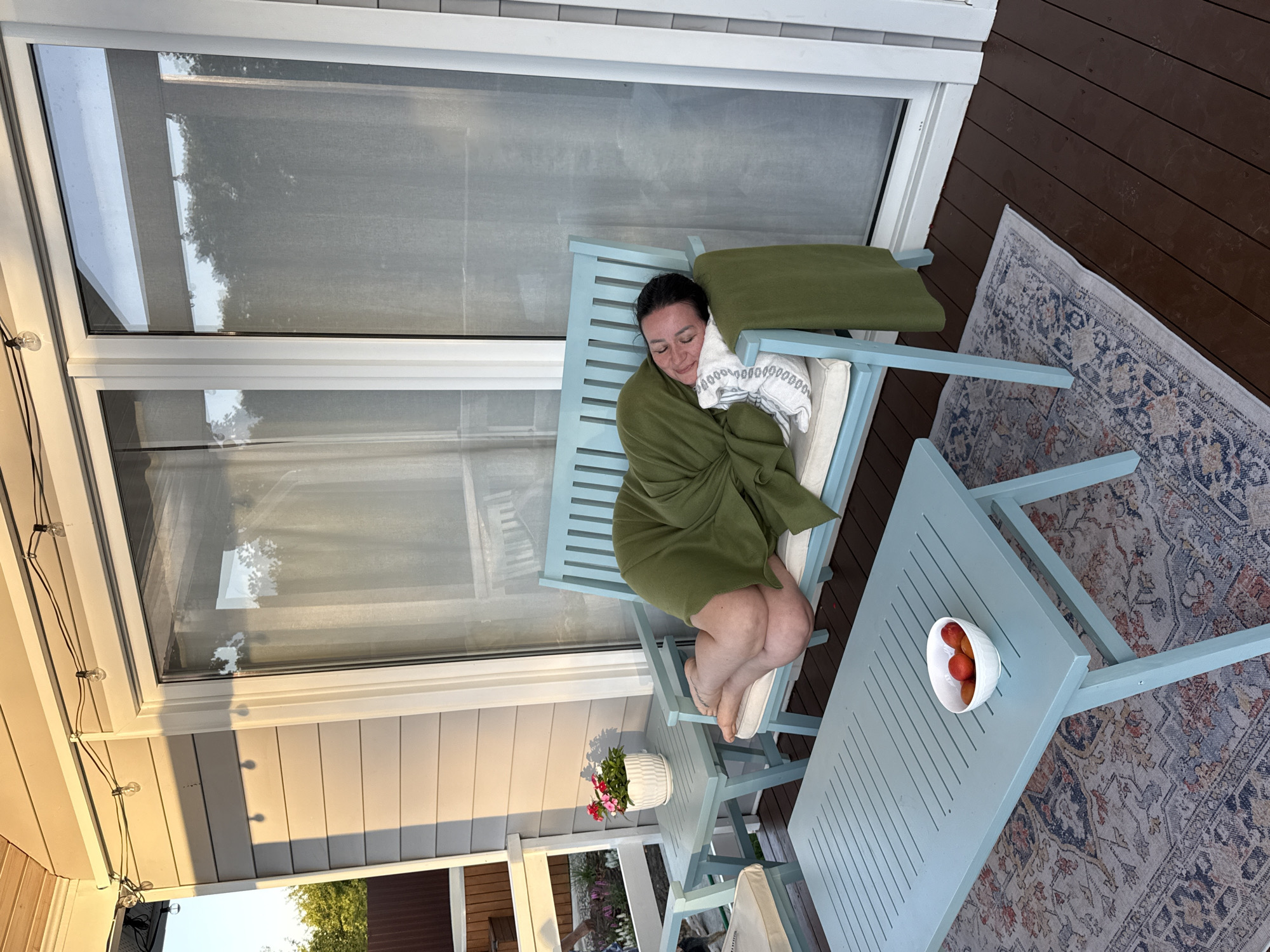 A woman is sleeping on a light blue bench on a wooden deck. She is covered with a green blanket and has a patterned pillow under her head. The bench has cushions. In front of the bench is a light blue coffee table with a white bowl of red fruit. A patterned rug is on the deck under the table and bench. A small side table with a potted plant is to the left of the bench. In the background are large glass doors and siding of a house. String lights are visible above the doors. The location is described as Starokorsunskaya, Russia.