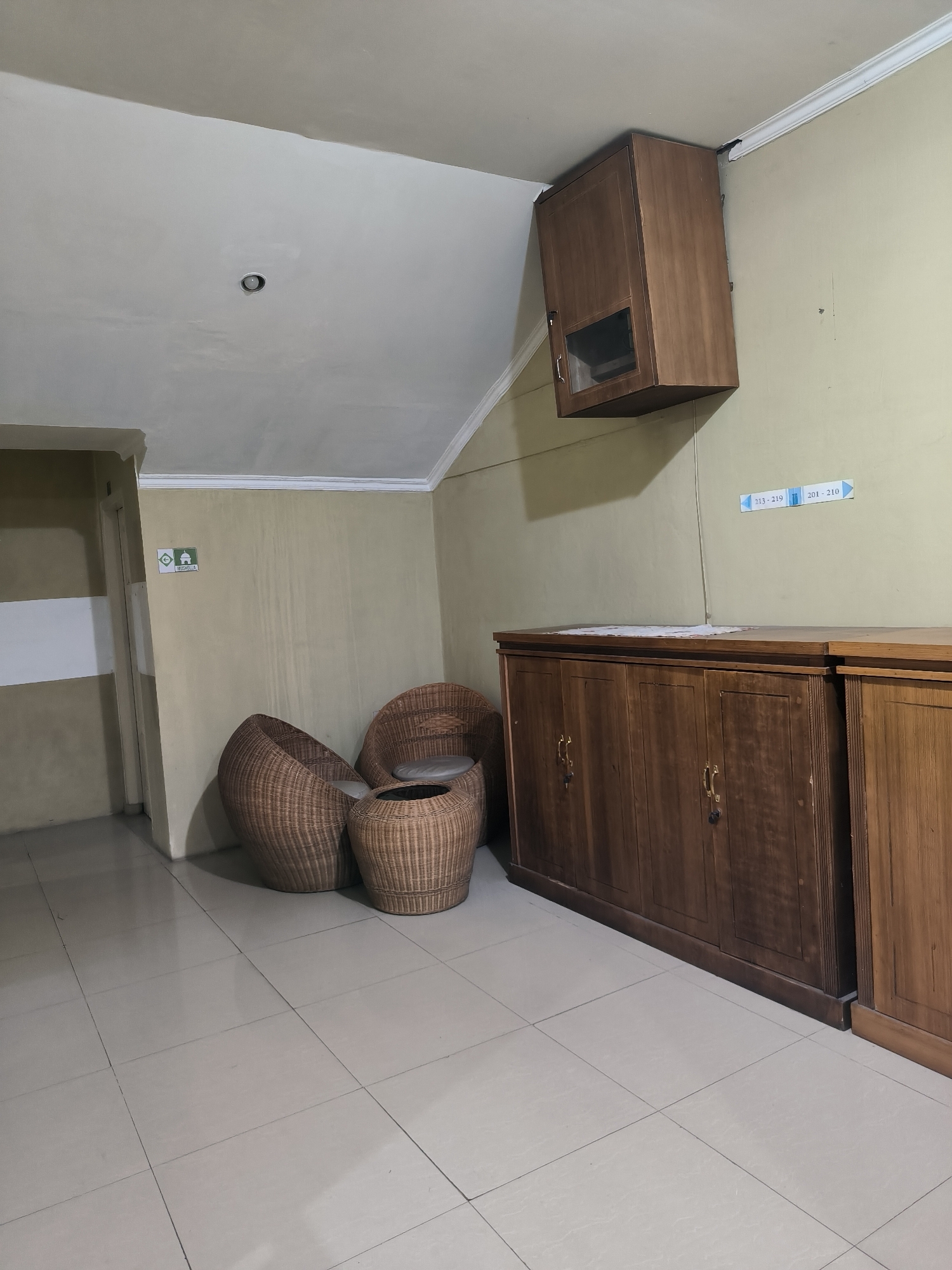 This media file captures an indoor waiting area or hallway, likely in a hotel or similar establishment, situated in Banda Aceh, Indonesia. The room features light beige-colored walls and a light-colored tiled floor, with a white ceiling that includes a single recessed downlight.
No people are visible, and there is no specific activity taking place, lending a quiet, unoccupied atmosphere to the scene.
Notable objects include a seating arrangement of two dark brown woven rattan armchairs with light-colored cushions, accompanied by a smaller, matching rattan stool or low table. Along the right wall, two long, dark wooden low cabinets with multiple doors and brass handles provide storage. Above these cabinets, a smaller, dark wooden wall-mounted cabinet hangs from the upper right corner, appearing slightly askew.
Visible text on the walls provides location cues:
* Near a doorway on the left wall, a green sign features white pictograms of a male figure, a female figure, and a mosque/prayer dome, with the word "MUSHOLLA" (prayer room) written below.
* On the right wall, above the lower cabinets, two small blue and white directional signs are present, indicating "213 - 219" and "201 - 210", likely denoting room numbers or sections of the building.
The lighting appears to be artificial, suggesting the image could have been taken at any time of day, with no natural light sources prominently visible. The overall impression is one of a functional, somewhat spartan waiting or transit area.