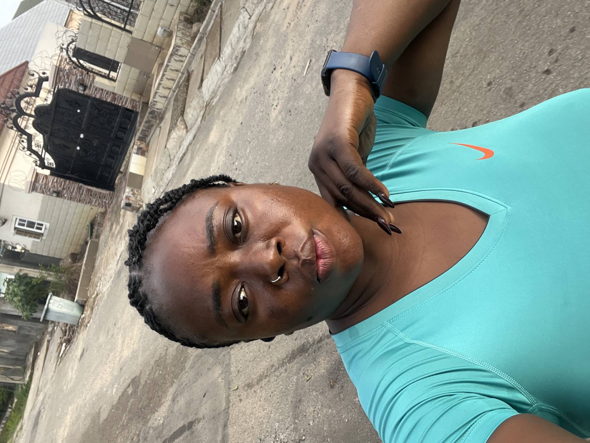 A selfie taken by a Black woman with braided hair and a nose ring, wearing a turquoise Nike shirt and a blue smartwatch. She is standing in an outdoor urban setting, possibly a street in Abuja, Nigeria, with a residential building and an ornate gate visible in the background. The woman is looking directly at the camera, with her right hand resting on her chin. The overall lighting suggests it is daytime, possibly overcast.