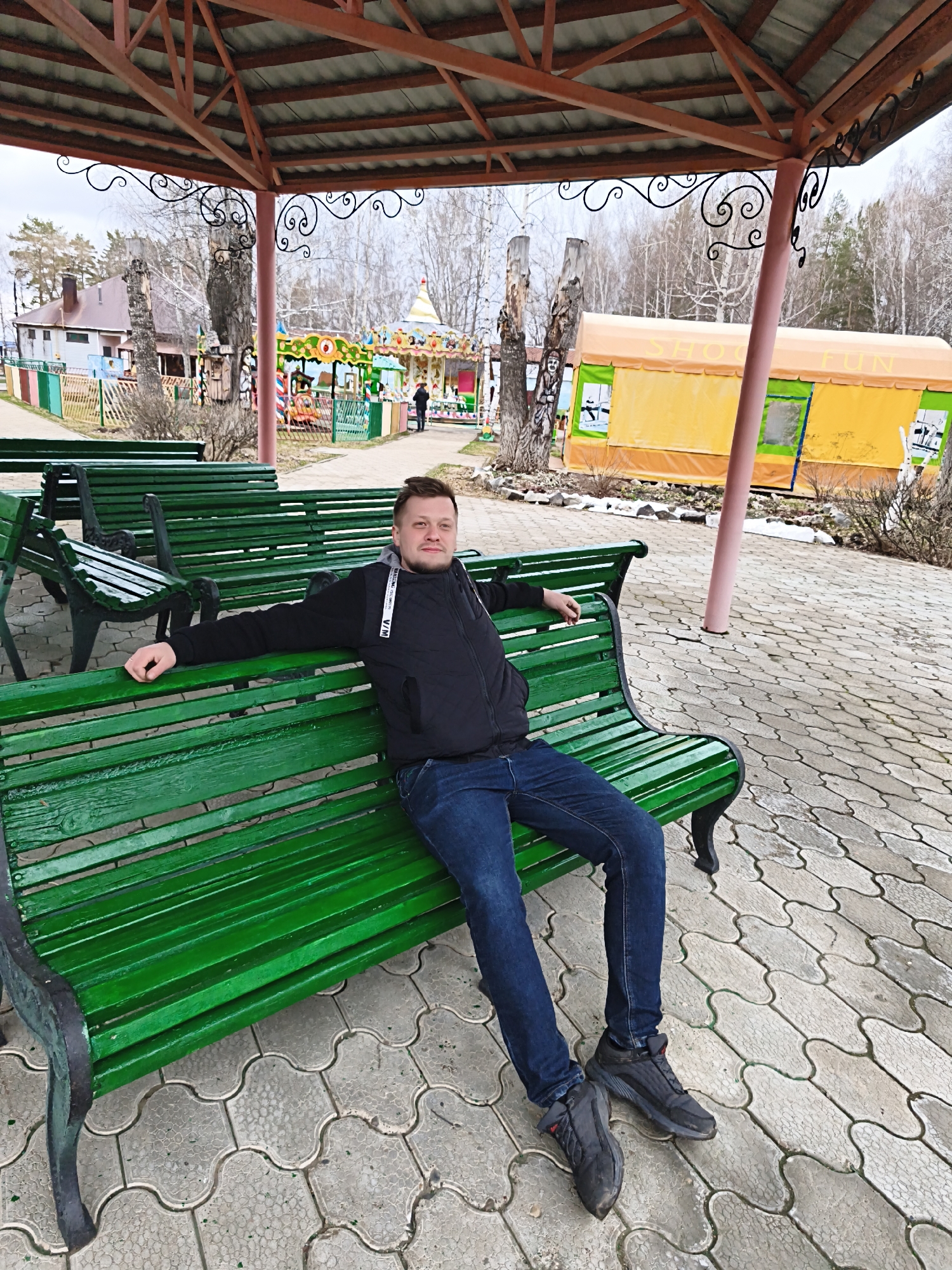 A man sits on a green wooden park bench under a sheltered pavilion in Chaikovsky, Russia, as shown by the "SHOOTING FUN" stand in the background. The man is wearing a black jacket, blue jeans, and dark athletic shoes. He is relaxed, with his arms stretched across the bench back. The ground is paved with interlocking stone blocks. In the background is a merry-go-round and various park structures. The environment suggests early spring, as the trees are bare.