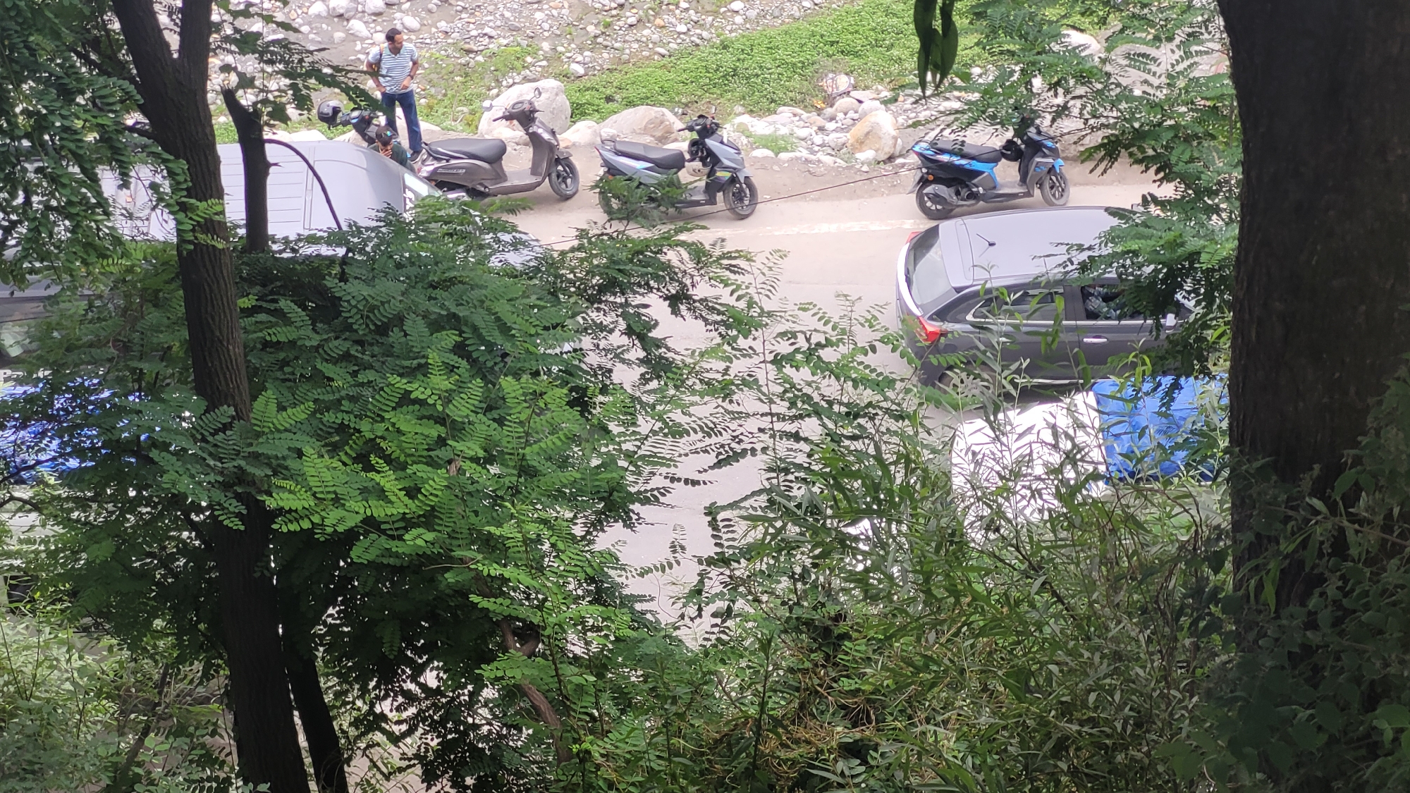 In Vashisht, India, a street scene unfolds with a focus on a road bordered by lush greenery and a rocky, elevated area. A man in a striped shirt and jeans stands near a white vehicle, facing away from the camera. Several scooters are parked or in motion on the asphalt road. A grey car is parked on the right side of the road, partially obscured by foliage. The environment suggests an outdoor, possibly rural or semi-urban setting, with natural elements like trees and rocks prominent. The lighting indicates it is daytime.