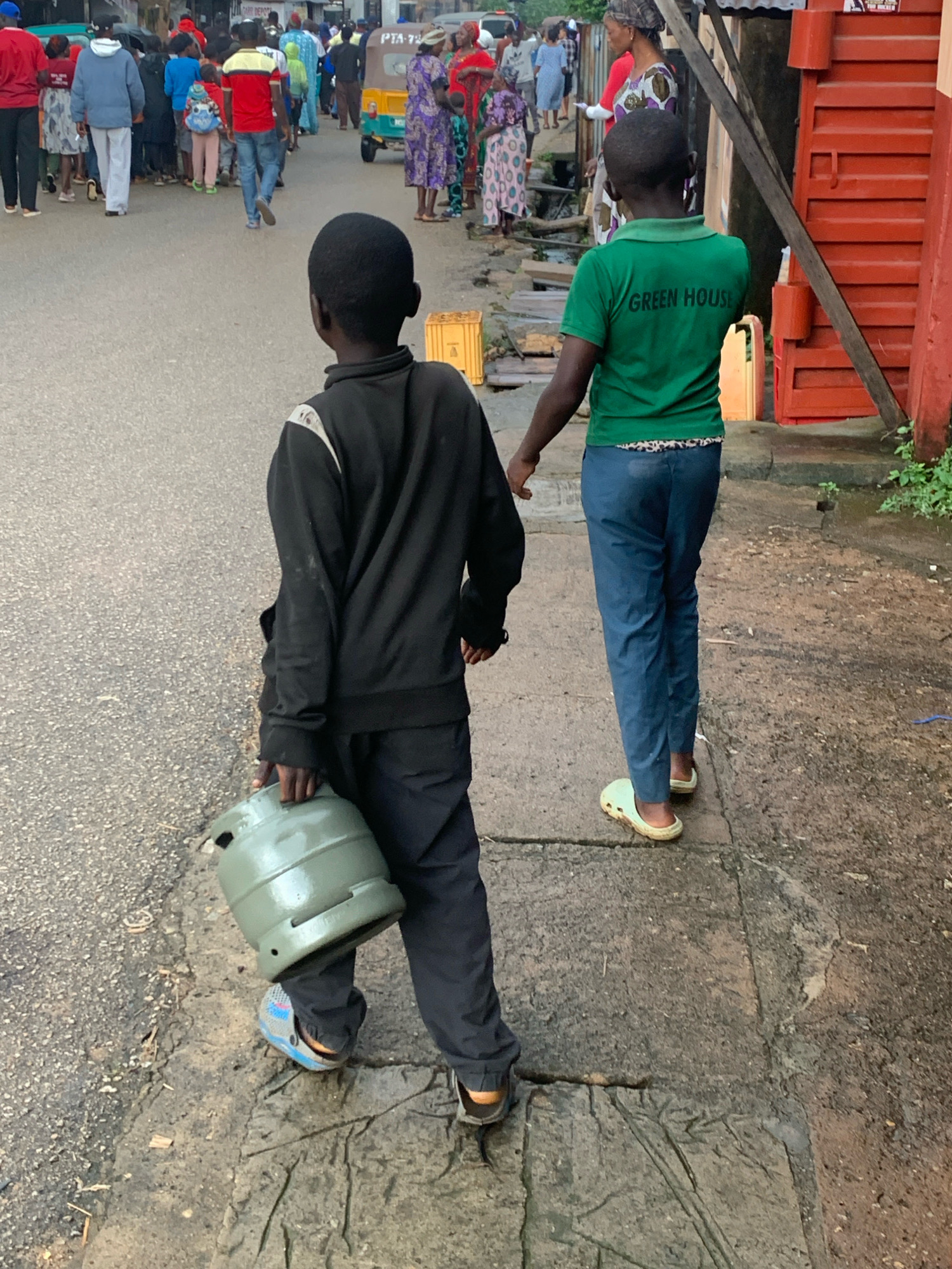Two young boys walk along a paved street in Jos, Nigeria. The boy on the left, viewed from behind, wears a black tracksuit and carries a green gas cylinder. He has on blue and grey croc-style shoes. The boy on the right, also viewed from behind, wears a green t-shirt with "GREEN HOUSE" printed on the back and blue trousers. He has on light-colored croc-style shoes. They are walking on a sidewalk next to a road that has other people walking on it. In the background, there is a red building with metal shutters.