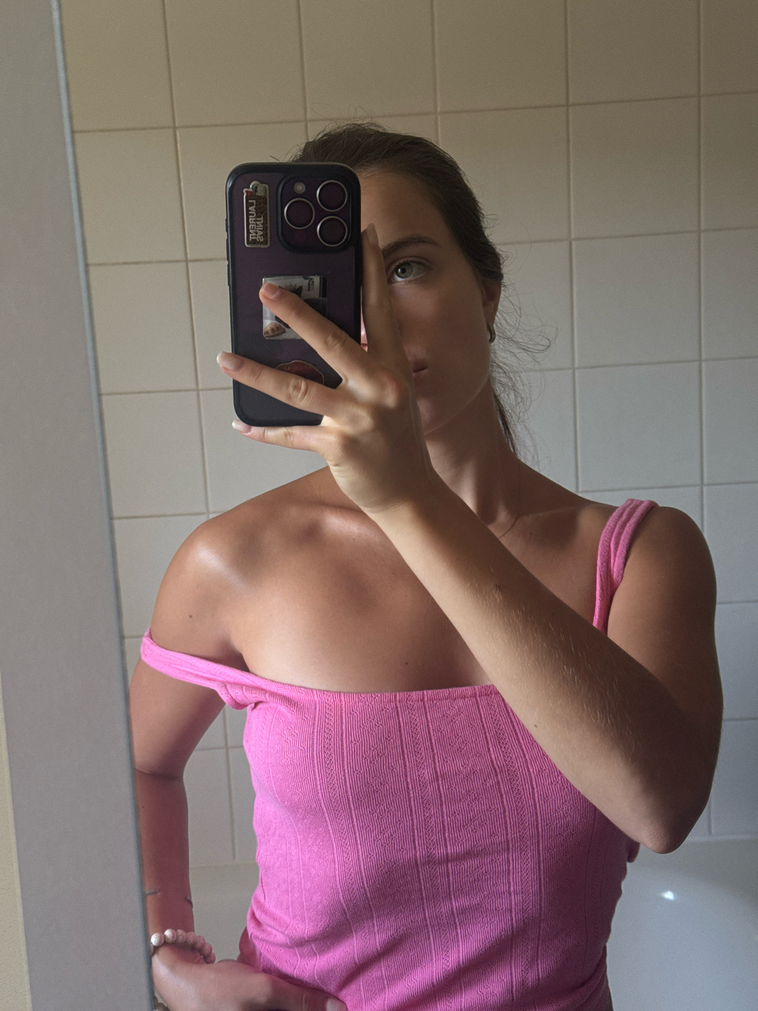 A young woman is taking a mirror selfie in a bathroom. She is wearing a pink ribbed tank top that is slightly off her shoulder. Her hair is pulled back in a ponytail, and she is holding a dark-colored phone in her right hand, obscuring the top half of her face. The bathroom has white tiled walls and a bathtub visible in the background. The lighting suggests it is daytime, possibly with sunlight entering the room. There are no other people visible in the image, and no context is provided about the city of Vienne, France.