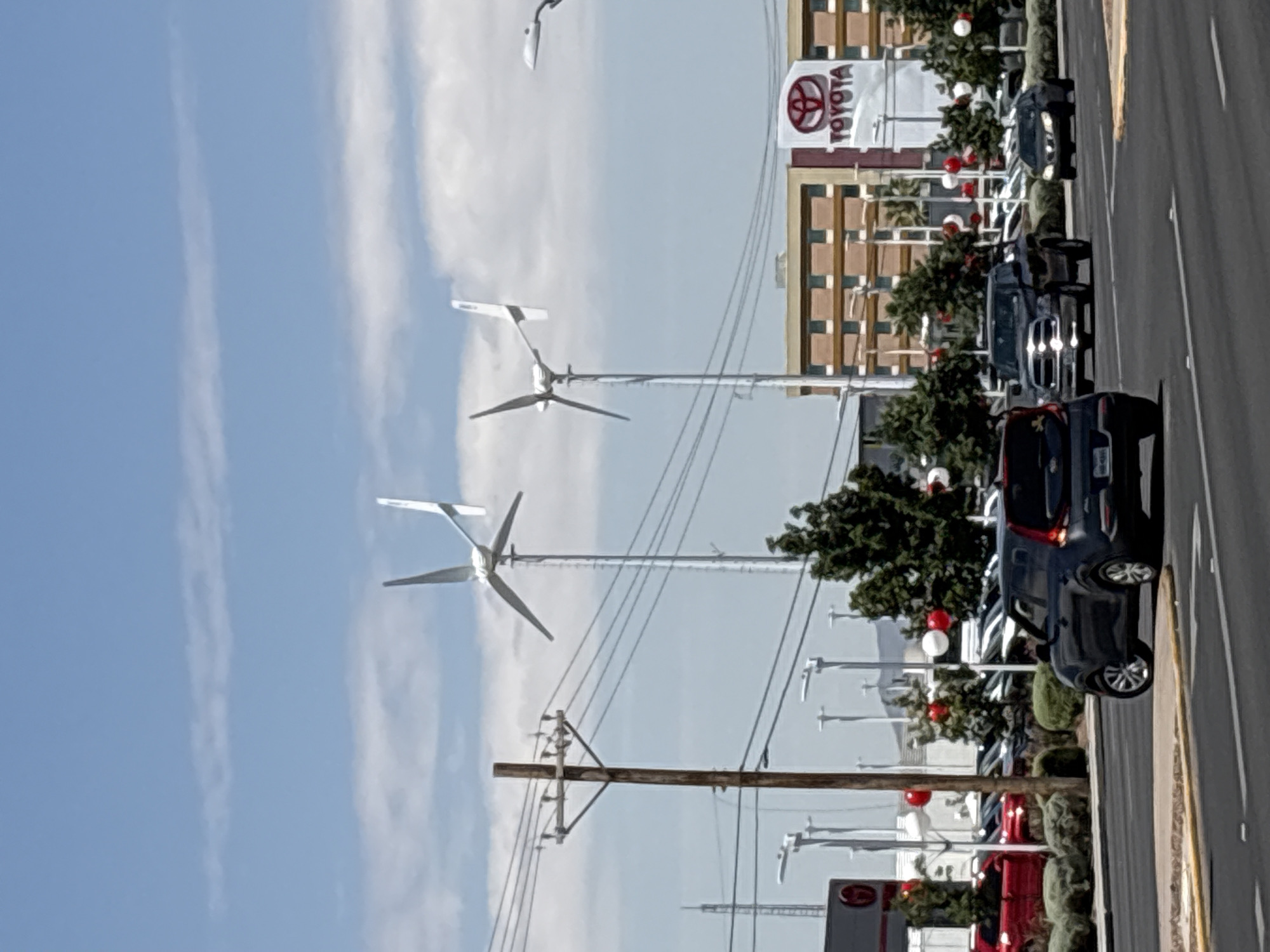 The image captures a street-level view of a Toyota dealership in El Paso, United States of America, under a partially cloudy sky. Two wind turbines stand prominently in the mid-ground, positioned behind a utility pole with power lines. The dealership building features a large Toyota logo and is adorned with red and white balloons, suggesting a special event or sale. Several cars are visible on the dealership lot, with some cars also driving on the road in the foreground. The overall scene depicts a commercial area with elements of renewable energy infrastructure integrated into the urban landscape.