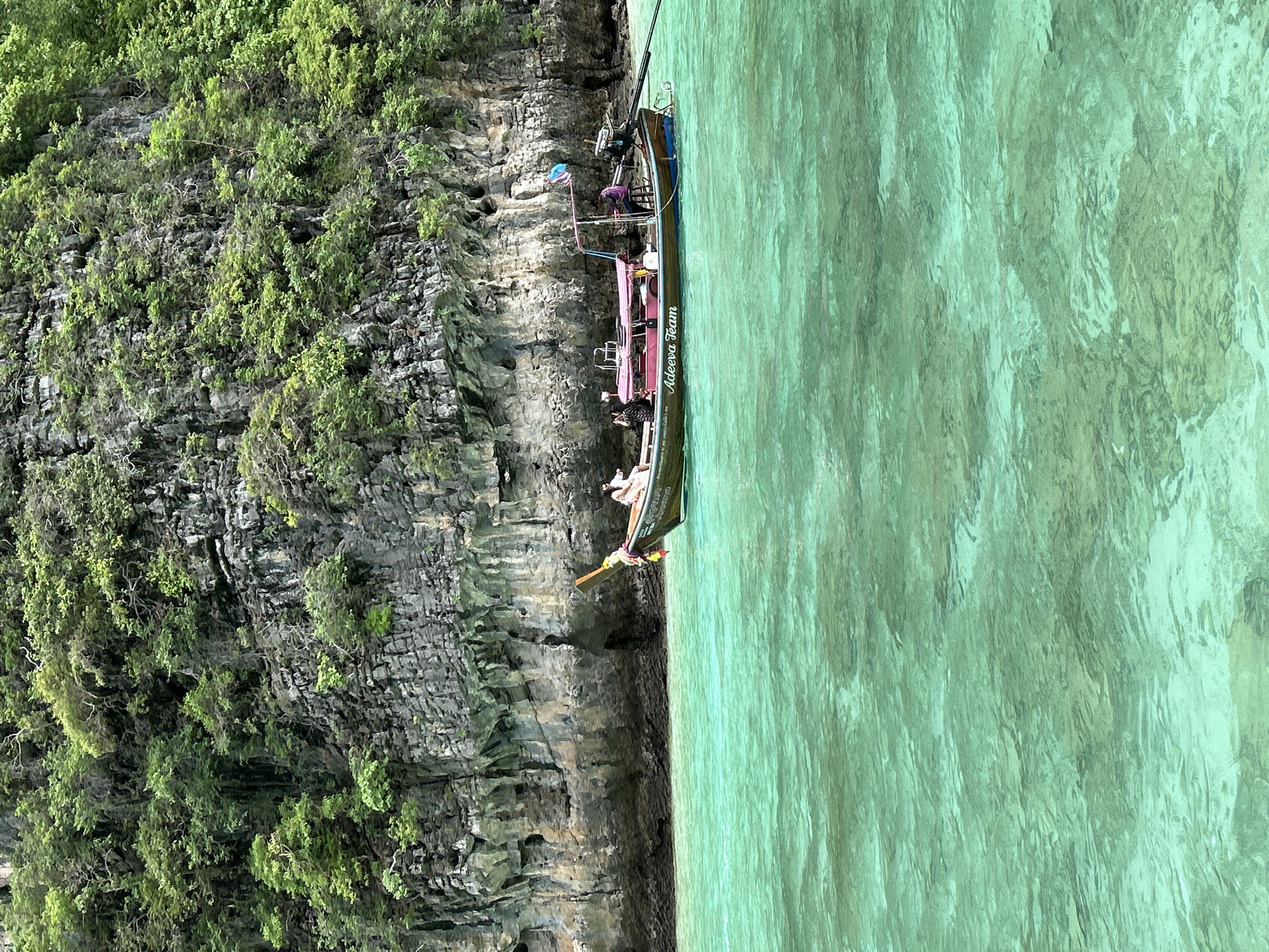 This media file captures a long-tail boat, a traditional Thai vessel, docked alongside a rocky, verdant cliff face. The water is a clear, vibrant turquoise, typical of tropical seas. The boat appears to be carrying passengers, with several individuals visible on board, some seated and others possibly standing. One person is leaning over the side of the boat, perhaps preparing to disembark or engage in an activity. The cliff is densely covered in lush green vegetation, suggesting a humid, tropical climate.
On the side of the boat, text is visible: "Adeeva Team". The overall scene evokes a sense of leisure and exploration, likely a tourist excursion in a scenic coastal area. The lighting suggests it is daytime, with natural sunlight illuminating the scene. The clarity of the water and the vibrant colors of the vegetation point towards fair weather. The rocky cliffs and lush foliage are characteristic of locations like Ao Nang in Thailand.