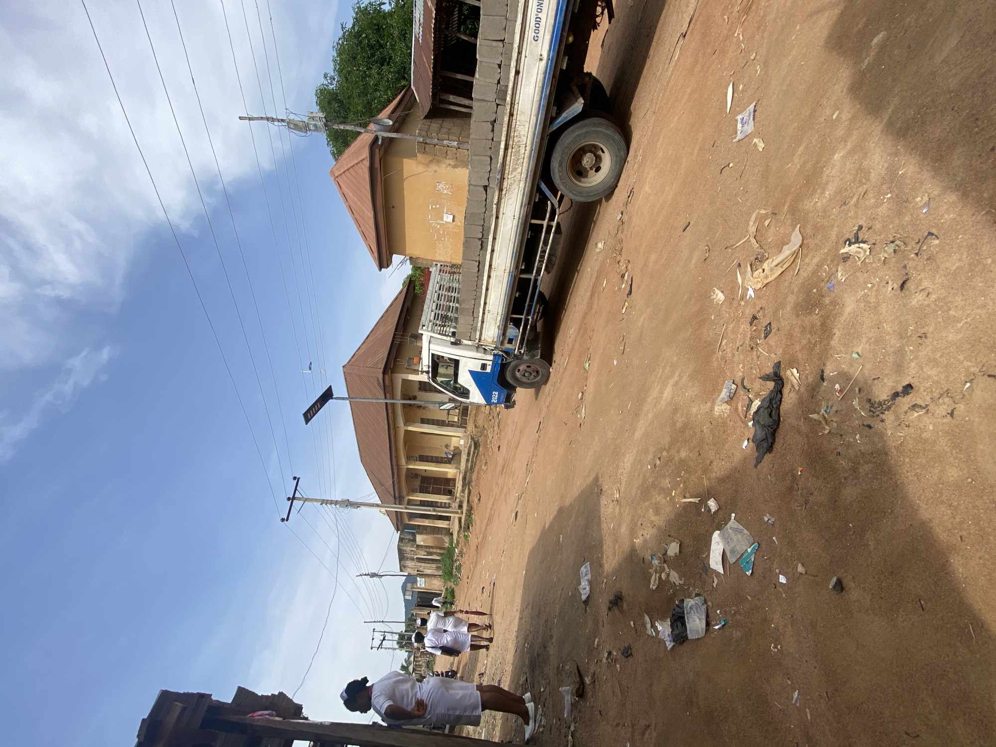 In Kuson, Nigeria, a street scene unfolds under a clear sky. A red dirt road, littered with plastic bags, paper scraps, and other debris, dominates the foreground. To the left, a woman in a white nurse's uniform stands near a wooden structure, her back mostly to the camera. Further down the road, two other individuals in similar white uniforms walk away from the viewer, one carrying a red umbrella. A white truck with a blue stripe, loaded with grey cinder blocks, is parked on the right side of the road, facing away from the viewer. In the background, a row of buildings with orange-tiled roofs line the street. Power lines and utility poles crisscross the sky. The overall impression is of a dusty, lived-in urban environment.