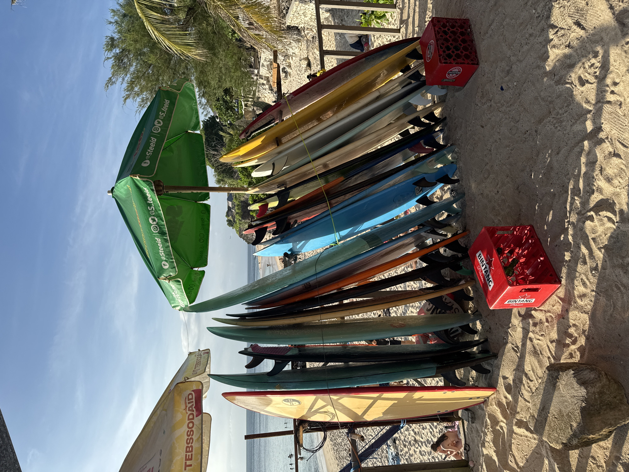 The image depicts a surfboard rack on a sandy beach in Kuta Selatan, Indonesia. The rack contains numerous surfboards of varying sizes and colors. Two red plastic crates, labeled "BINTANG Pilsener," are positioned near the rack. Several beach umbrellas are visible, including one with the text "TEBSSODAID" and another with social media handles "@Steeid." The beach stretches towards the ocean, with people visible in the distance. Palm trees and other vegetation are present in the background. The sky is partly cloudy.