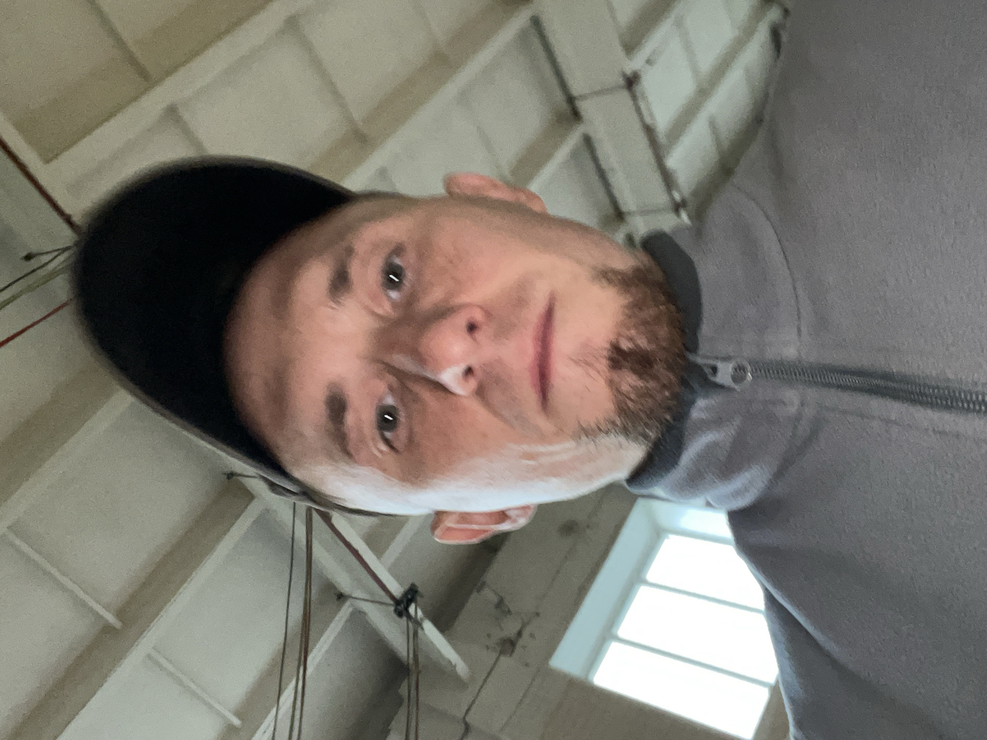 The media file is a close-up, low-angle selfie of a man wearing a grey zip-up hoodie and a dark baseball cap. The man has light skin, a beard, and is looking slightly upwards and to the left. His eyes are open and he appears to be in a well-lit indoor environment. The background shows a white ceiling with visible beams and light fixtures, suggestive of an industrial or warehouse-like setting. There are also windows in the background, which are brightly lit, indicating it is daytime. The image is rotated, with the man's head appearing at the top of the frame and the background elements below. No text is visible in the media file. The setting appears to be Sarapul, Russia.