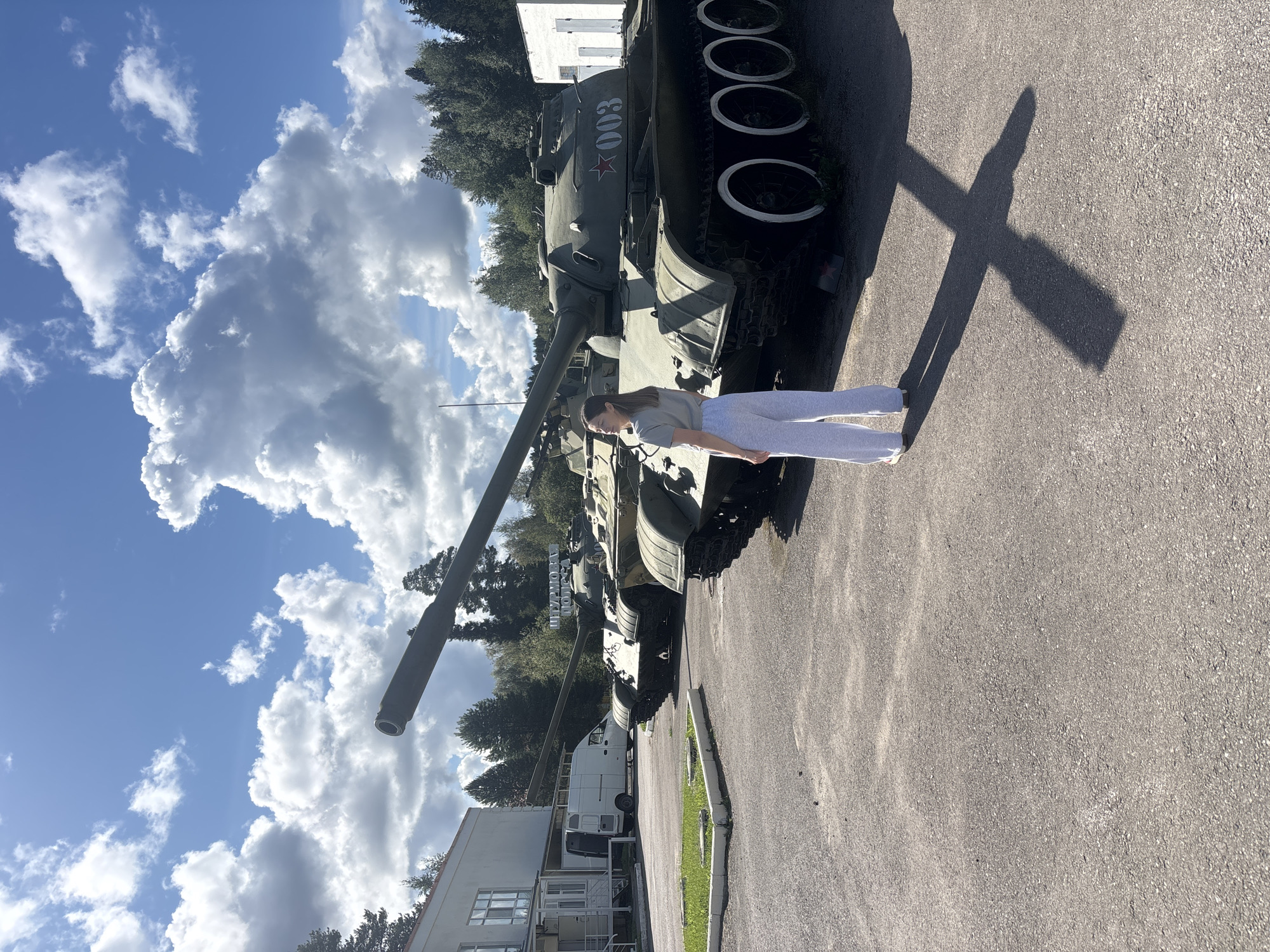 A woman stands in front of a row of olive-green tanks in what appears to be a military museum or outdoor exhibition. The tanks are lined up on an asphalt surface, with a grassy area and a white building to the left. The woman, wearing a grey t-shirt and light-colored sweatpants, is facing away from the camera, looking up at the sky. The tanks have distinctive markings, including a red star and the number "003" on the side of the closest one. The sky is blue with scattered white clouds, and trees are visible in the background. The location is identified as Gamy, Russia.