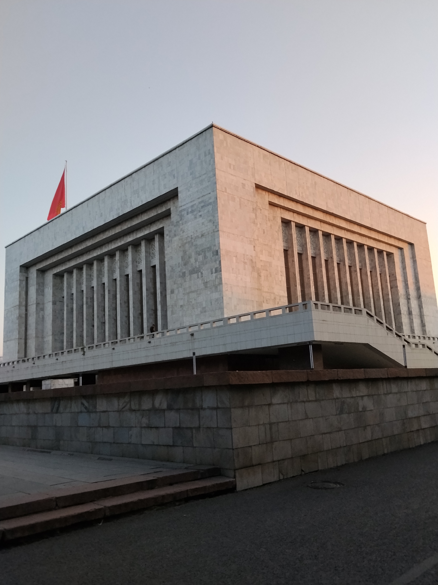 The image shows the State Historical Museum in Bishkek, Kyrgyzstan, under a pale sky. The museum is a large, imposing building with a facade made of light-colored stone, possibly marble, with a pattern of vertical indentations. A red flag flies from the top left corner of the building. In front of the museum is a raised platform with a white railing and a ramp leading up to the building. The platform is supported by a wall constructed of large, gray stone blocks. The ground in front of the wall is paved with asphalt. Two people are visible standing on the platform near the railing. The overall impression is one of grandeur and solidity, typical of Soviet-era architecture.