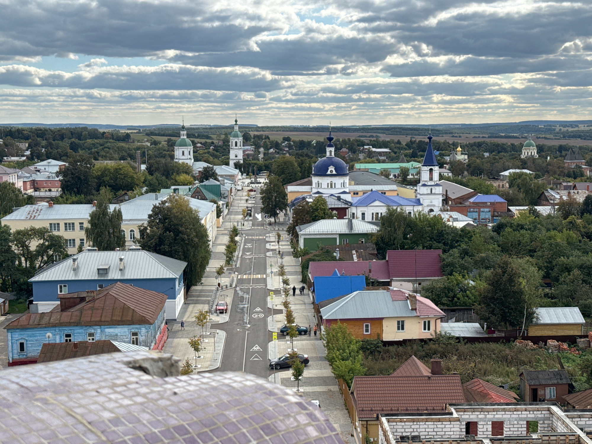 The image provides an aerial view of a city street in Zaraysk, Russia, with buildings and greenery. The street is paved and lined with trees and sidewalks. Several cars are parked along the street. The buildings are varied in style and color, with some featuring pitched roofs and others flat roofs. Church spires are visible in the distance, indicating a historical or religious context. The sky is partly cloudy, with a mix of gray and white clouds. The overall impression is of a moderately populated urban area with a mix of residential and commercial structures.